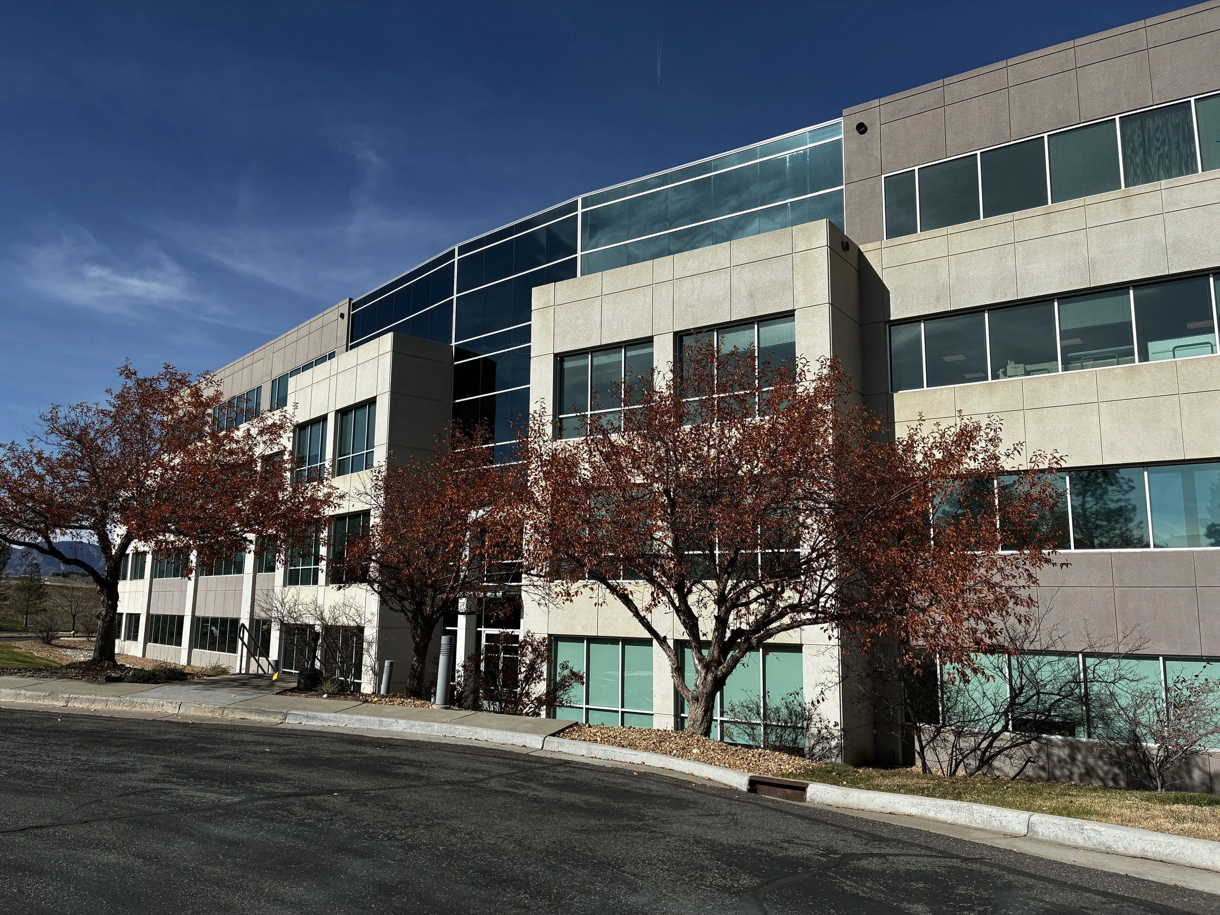 Modern office building with large glass windows and beige exterior. Trees with reddish-brown leaves in front and a paved parking area.