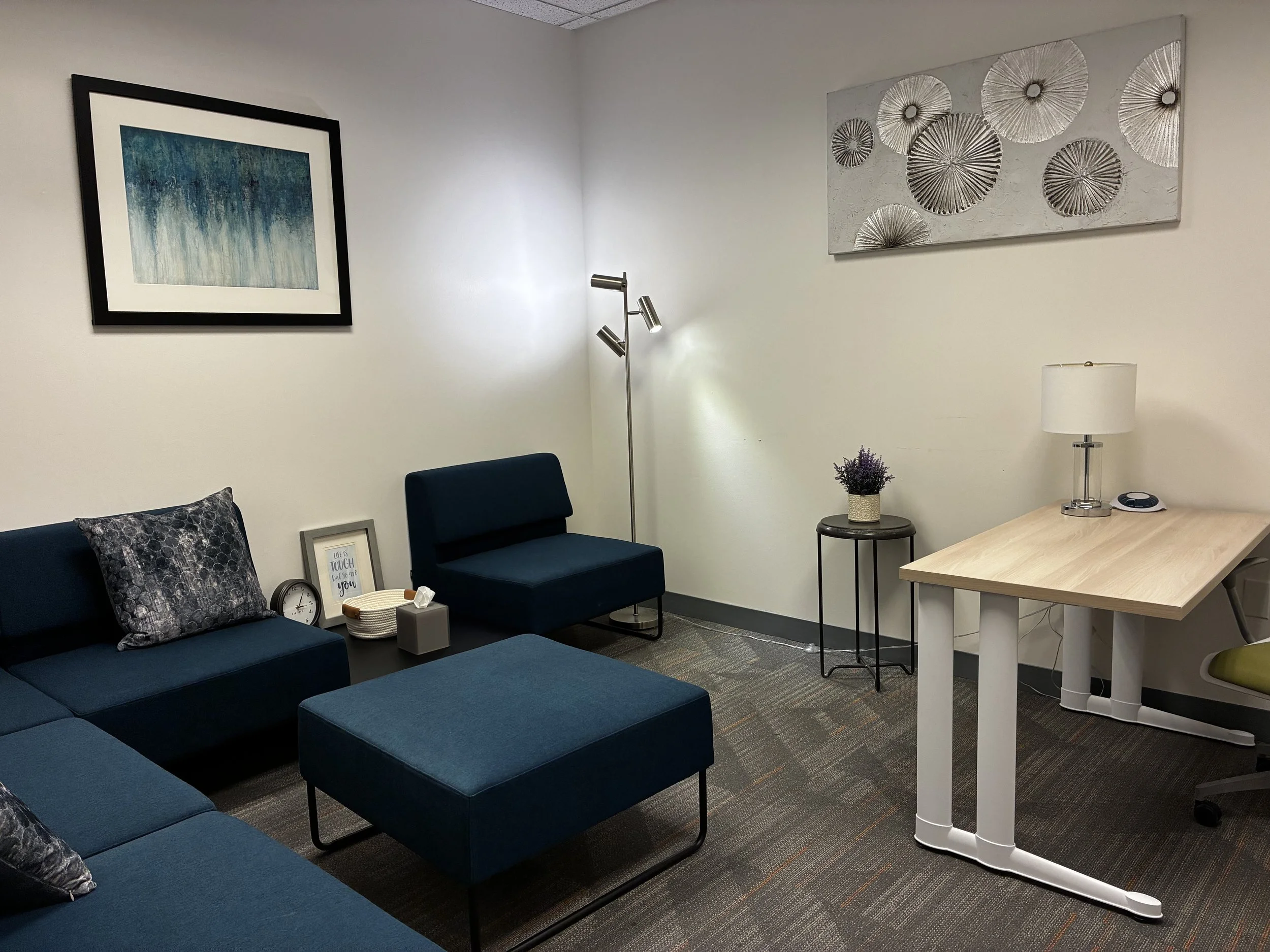 A living room with navy blue sectional sofa, a matching armchair, a ottoman, wall art, a wall lamp, a side table with a plant, a desk with a lamp, a phone, and a green office chair.