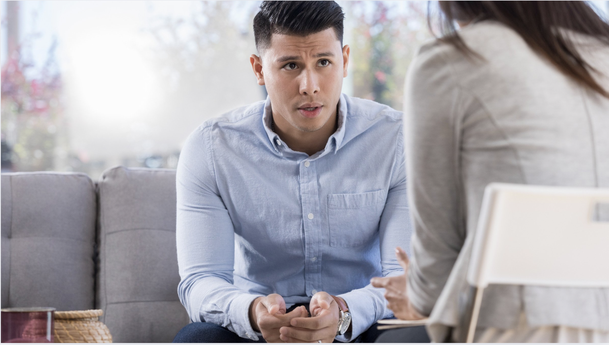 A young man in a light blue shirt looks concerned while talking with a woman in a beige blazer at a therapy session in a bright room.