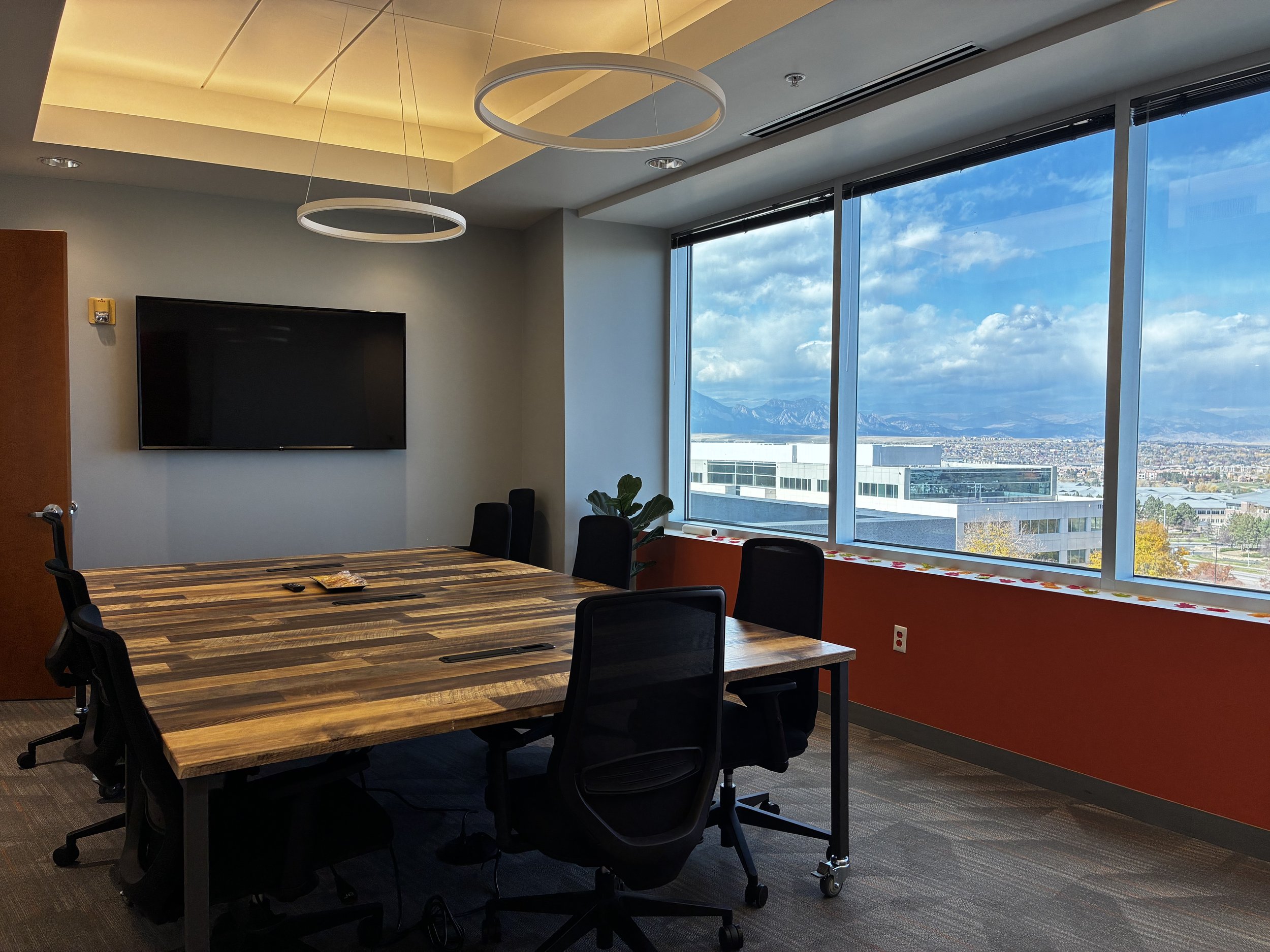 Conference room with a large wooden table, black chairs, a wall-mounted TV, and large windows with a cityscape and mountains in the background.