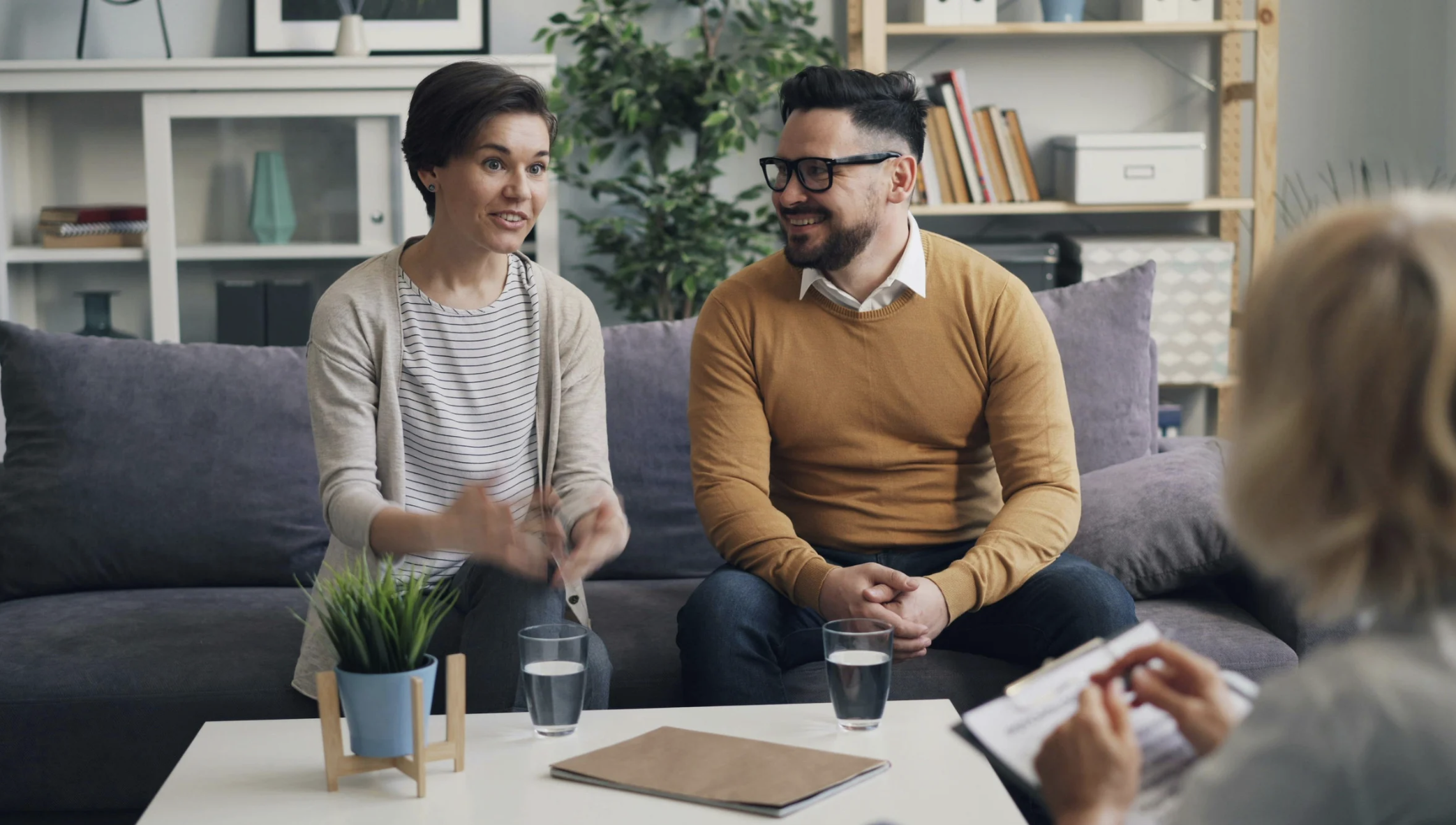 Two people, a woman and a man, sitting on a sofa in a living room, talking to a third person holding a notepad. There are two glasses of water and a potted plant on a coffee table.