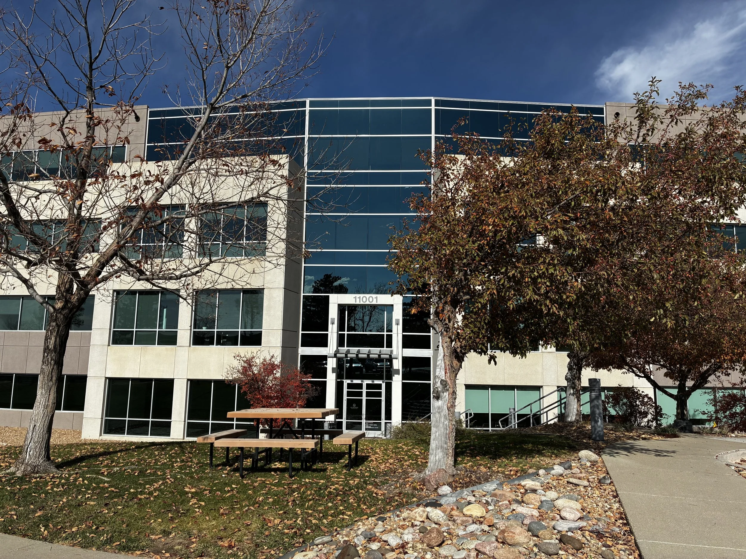 Front view of a modern office building with glass windows and an entrance, surrounded by trees with fall foliage, a sidewalk, and a picnic table outside.