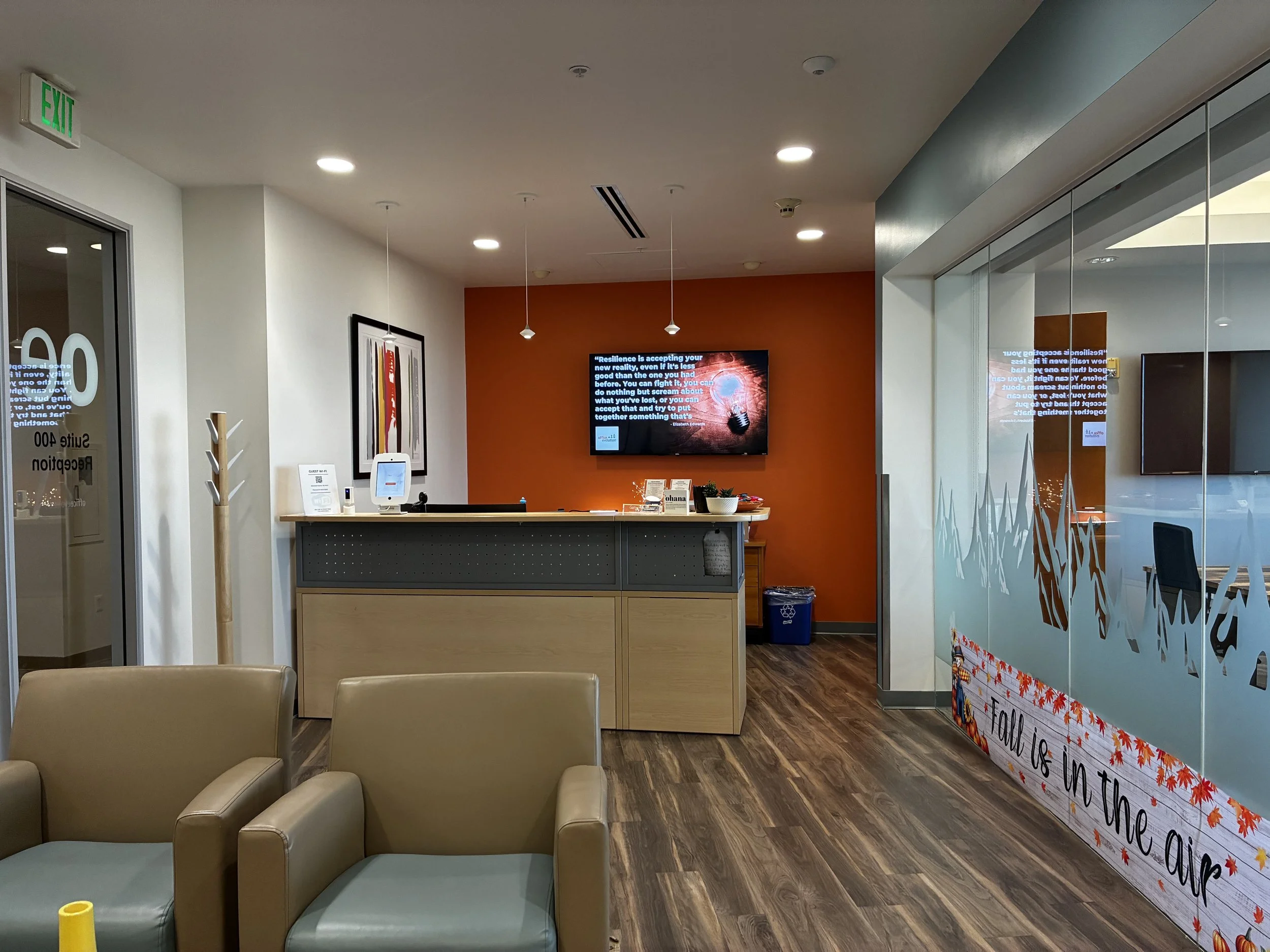 Lobby reception area with seating, a front desk, artwork on the wall, and an orange accent wall with a TV displaying a motivational message.