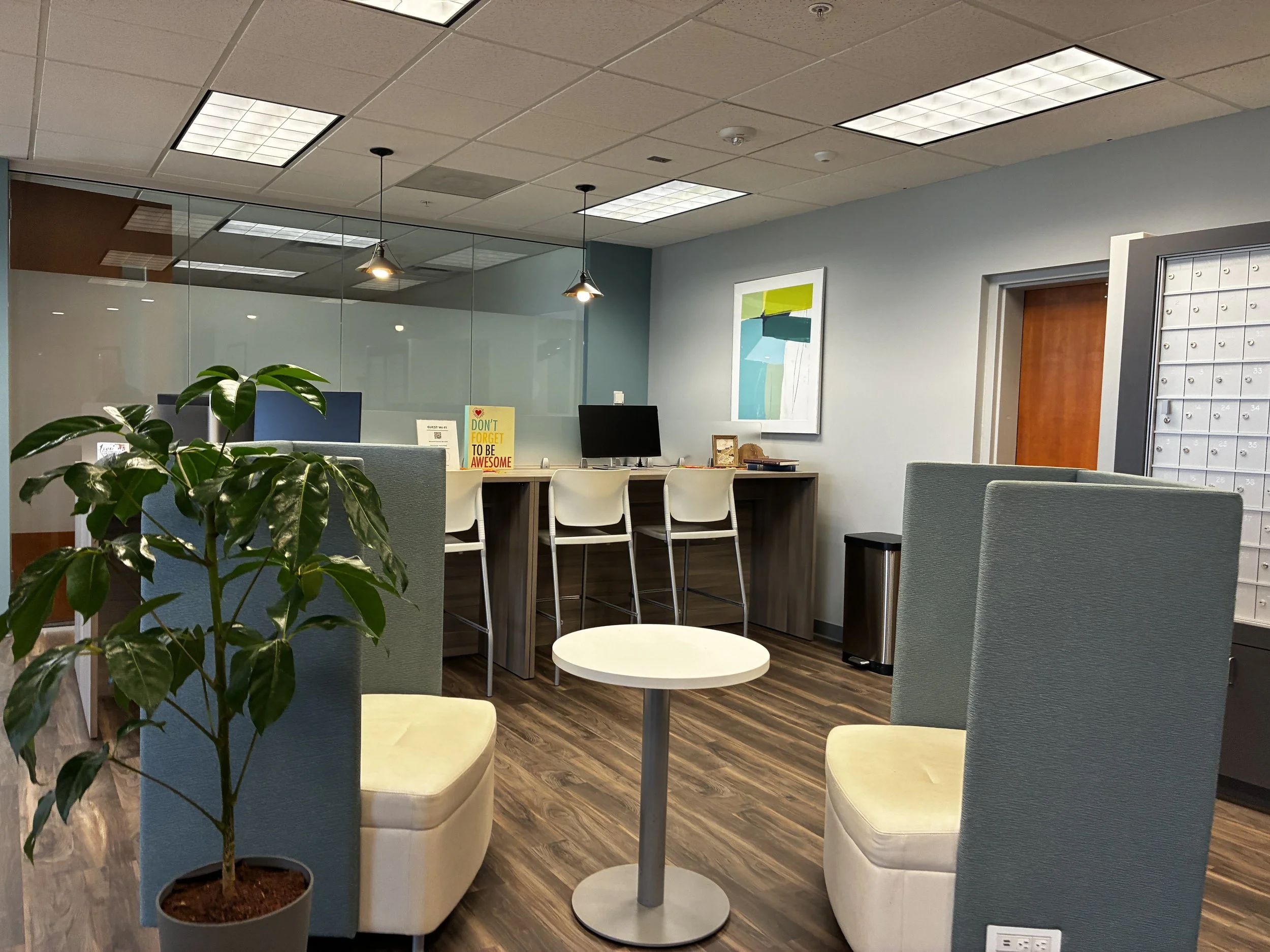 Modern office lobby with seating area, potted plant, small round table, and a reception desk with chairs, monitor, and decorative artwork on wall.