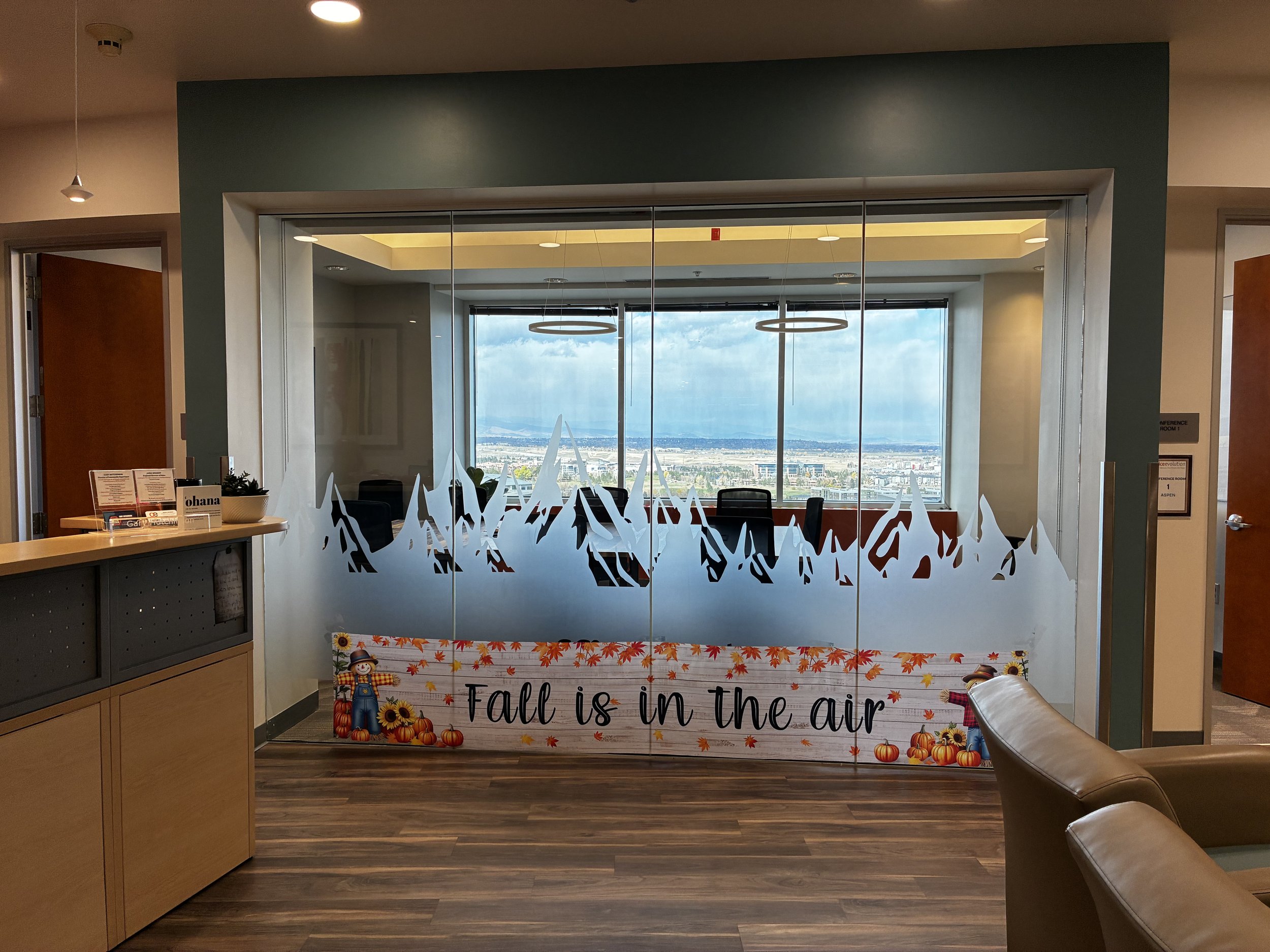 Interior view of an office with a glass wall decorated with fall-themed graphics and a sign that reads 'Fall is in the air,' featuring pumpkins, sunflowers, and autumn leaves, with a cityscape visible through large windows in the background.