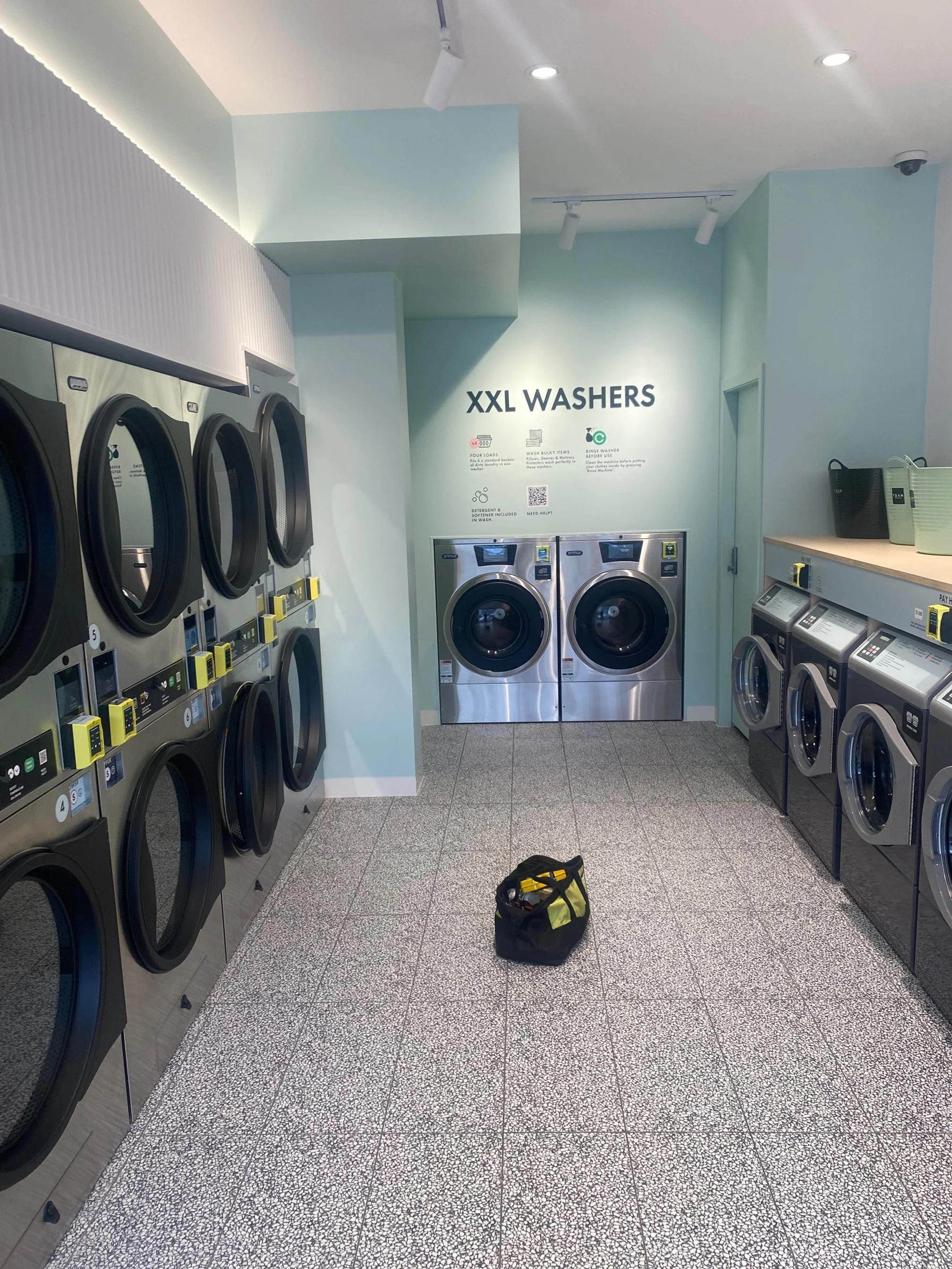 Laundry room with multiple washing machines, including large XXL washers, on both sides and a pair of front-loading washing machines at the far end, with a small bag on the floor in the center.