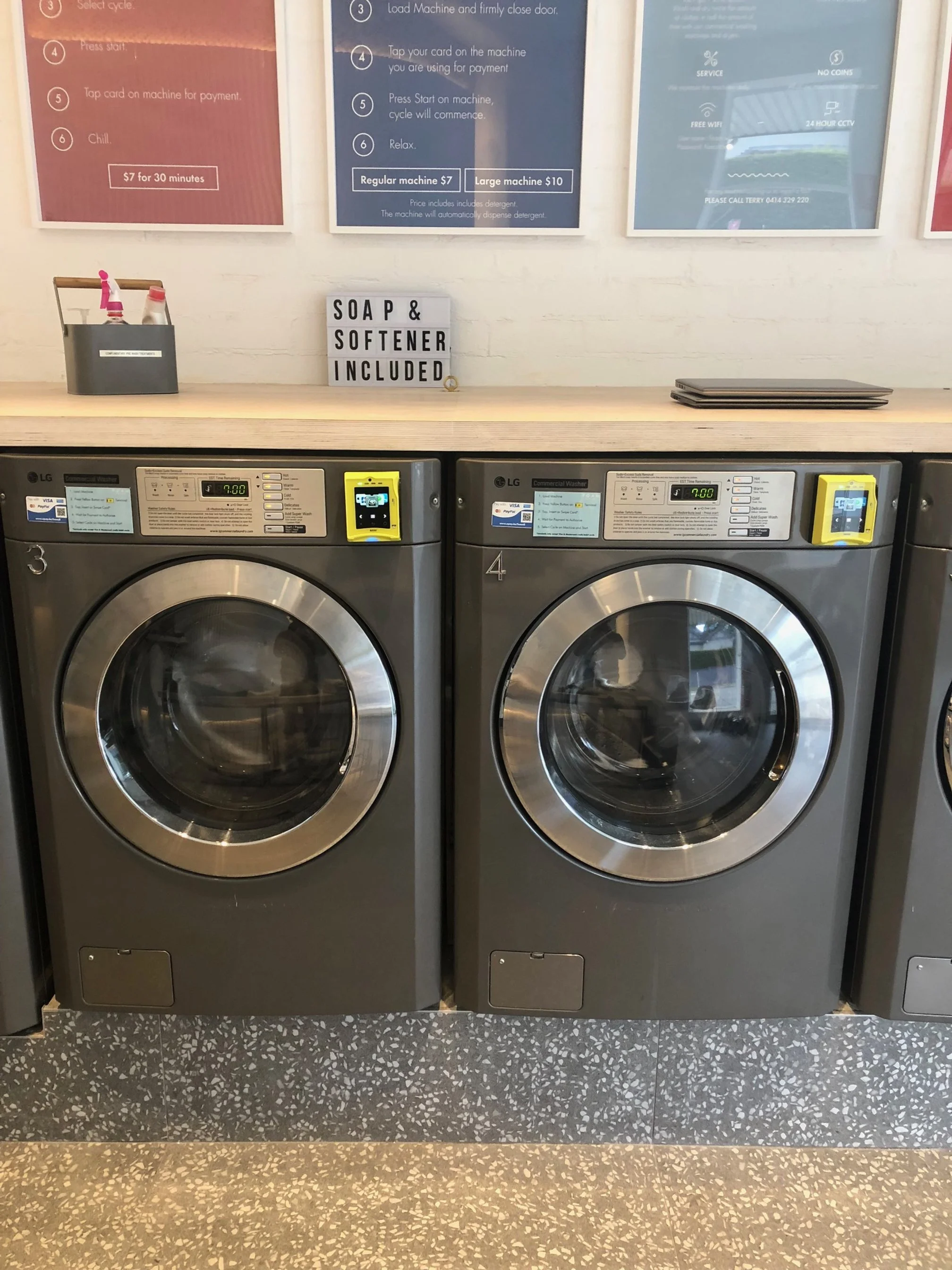 Two industrial washing machines in a laundromat, with a sign above stating 'Soap & Softener Included'.