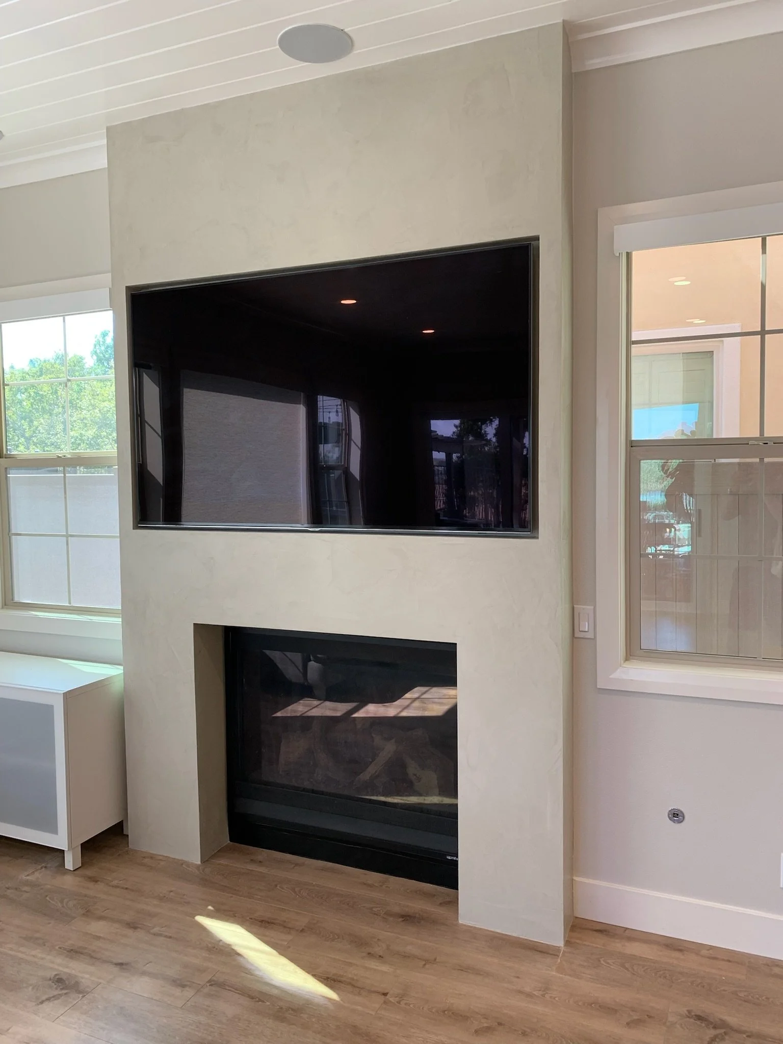 Living room with a wall-mounted TV above a fireplace, with large windows on either side allowing natural light.