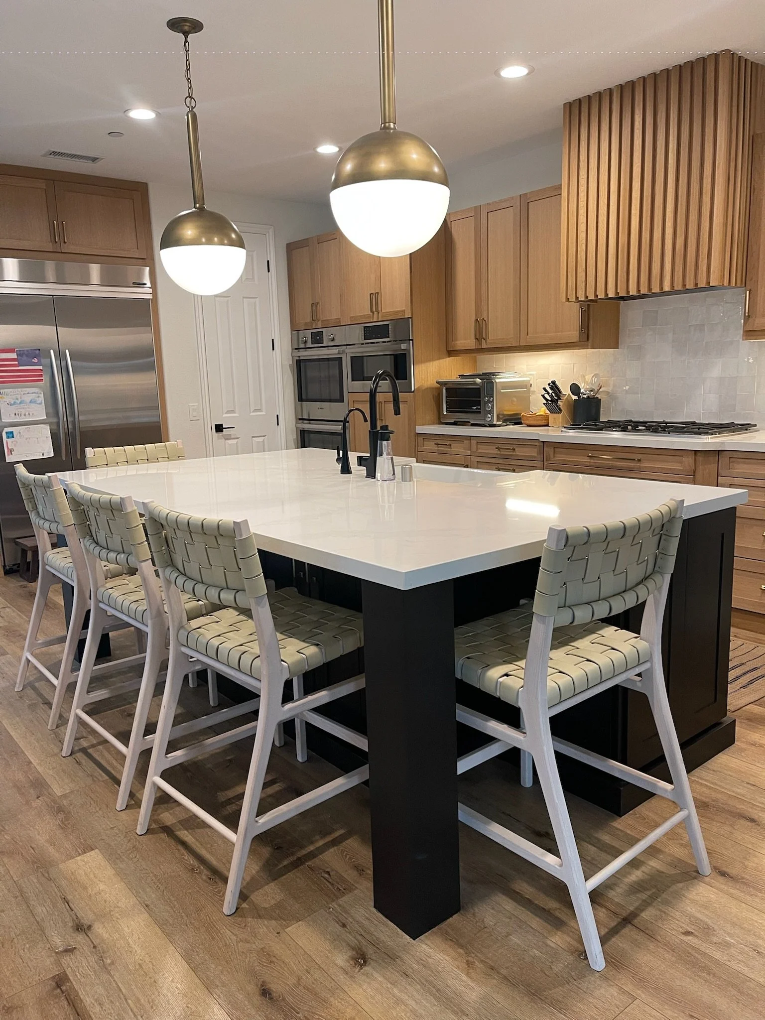 Modern kitchen with white island and six woven-back chairs, wooden cabinets, stainless steel appliances, and pendant lighting.