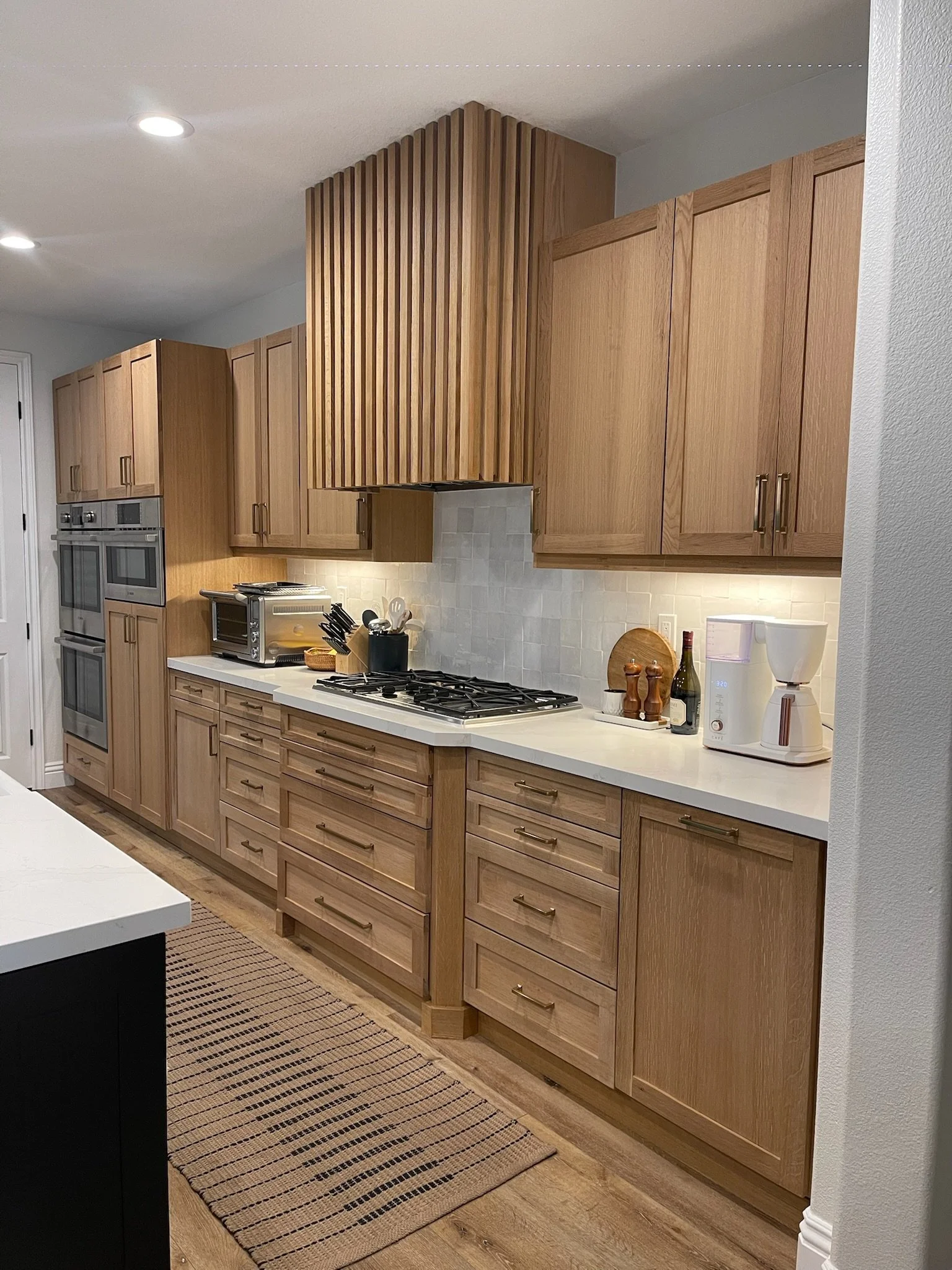 Kitchen with light wood cabinets, white countertops, built-in ovens, a stovetop, a toaster, a coffee maker, and decorative items such as wine bottles and wooden cutting boards.