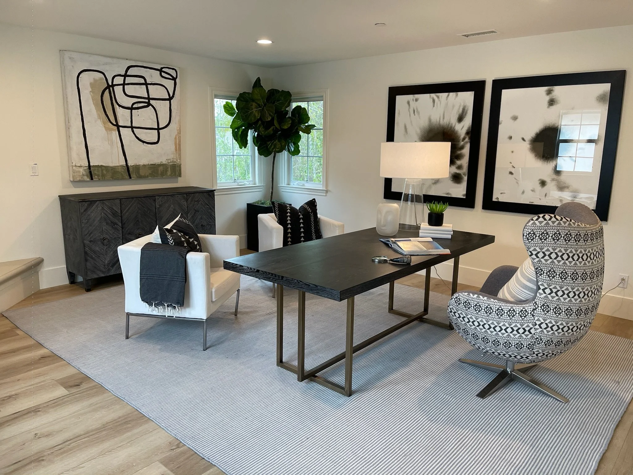 Modern living room with a black table, white armchairs, a patterned accent chair, abstract black and white wall art, and a tall green plant near the window.
