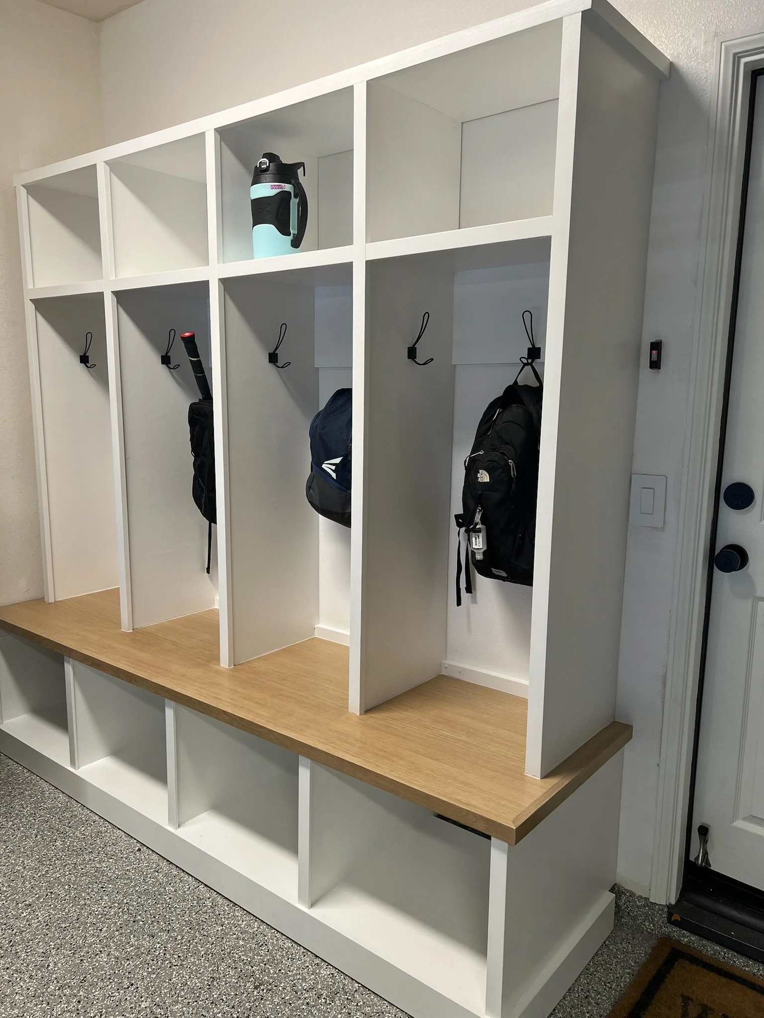 A white wooden cubby locker with four hanging lockers, some with backpacks, and an upper shelf holding a water bottle.
