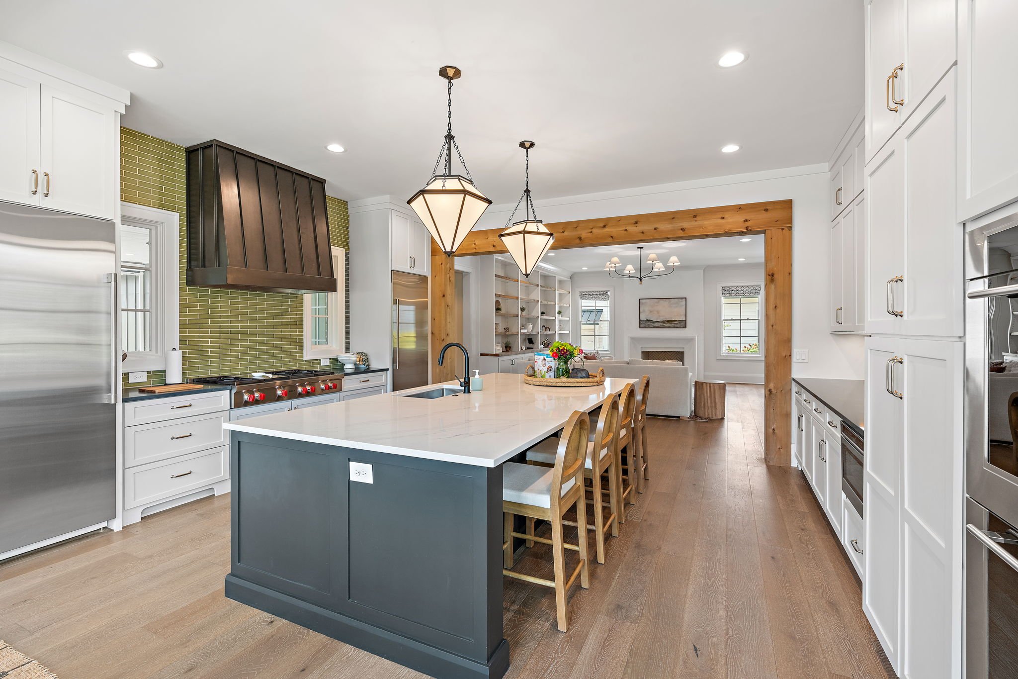 Modern kitchen with white cabinets, stainless steel appliances, a large island with a dark blue base, wooden chairs, and pendant lights, opening into a living room with a sofa, fireplace, and windows.