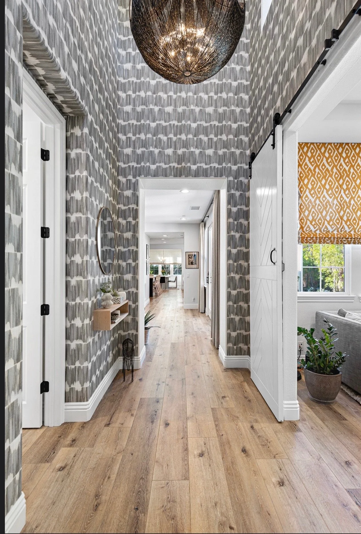 A hallway with patterned gray and white wallpaper, wood flooring, a circular mirror, and a small shelf with plants, leading to a dining area with windows and a living room with a gray couch and large window with orange patterned blinds.