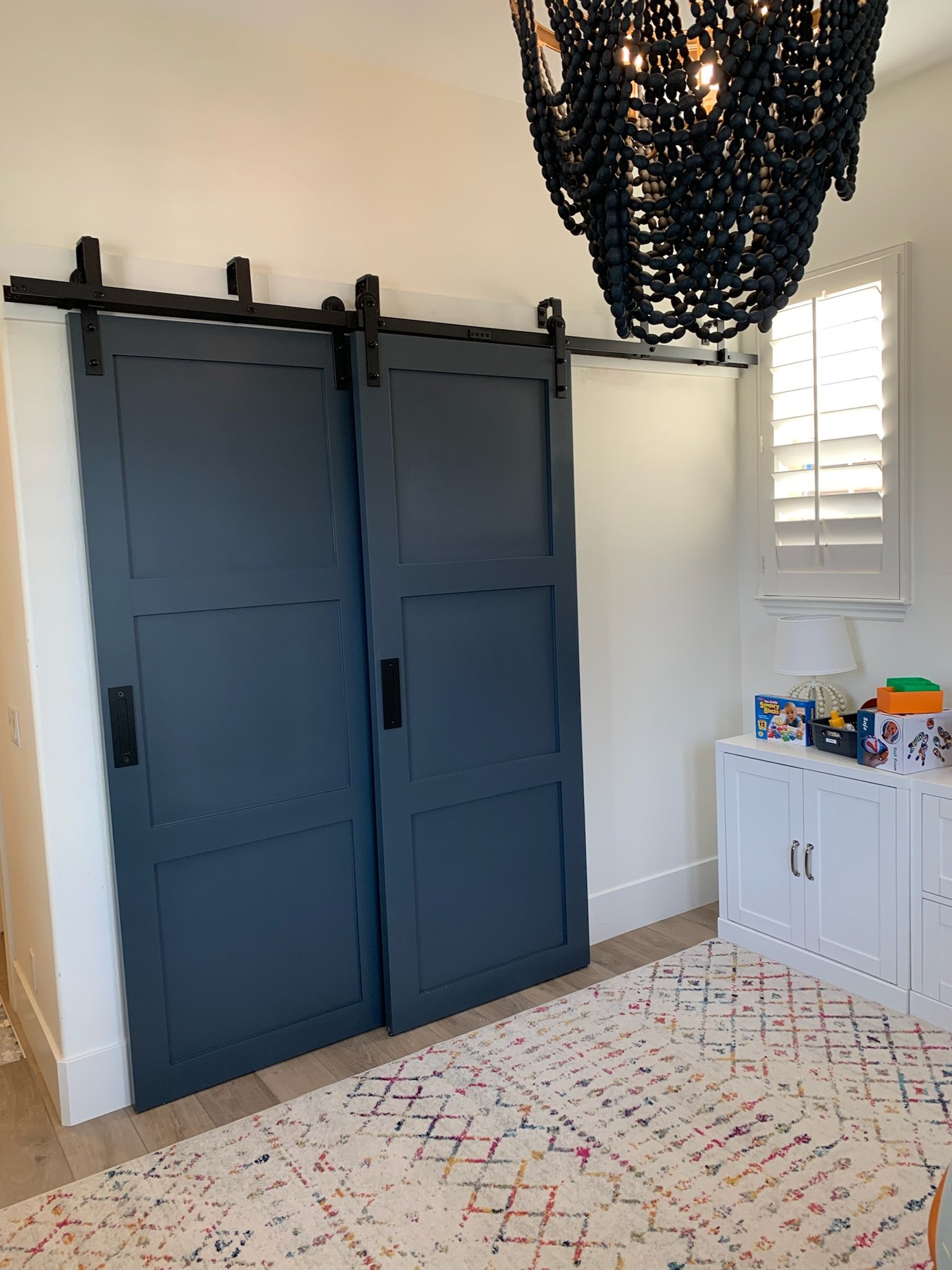 Blue sliding barn doors in a room with white walls, a white cabinet and a window with shutters, and a black beaded chandelier hanging from the ceiling.
