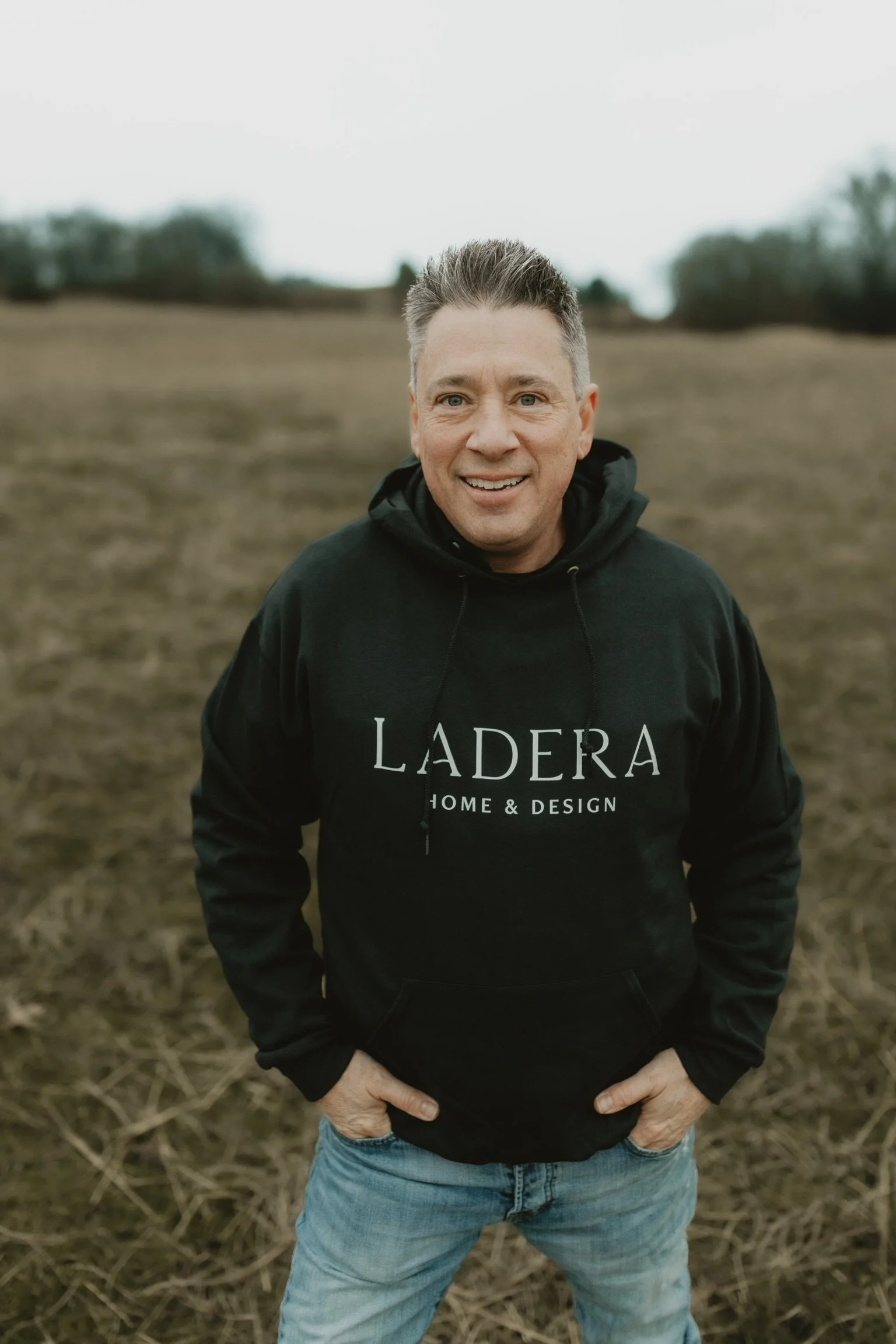 A smiling man with short gray hair standing outdoors in a field, wearing a black hoodie with "LADERA HOME & DESIGN" printed on it, and blue jeans.