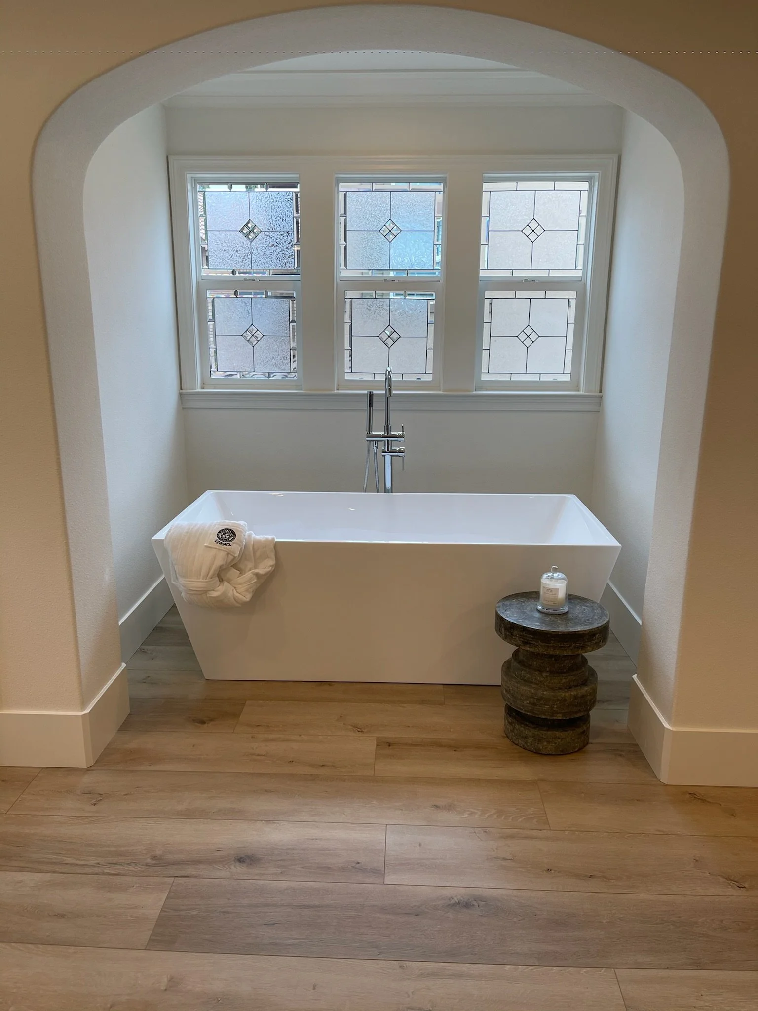 Bathroom with a white bathtub, a dark wooden side table, a bottle of soap, and four decorative windows with stained glass.
