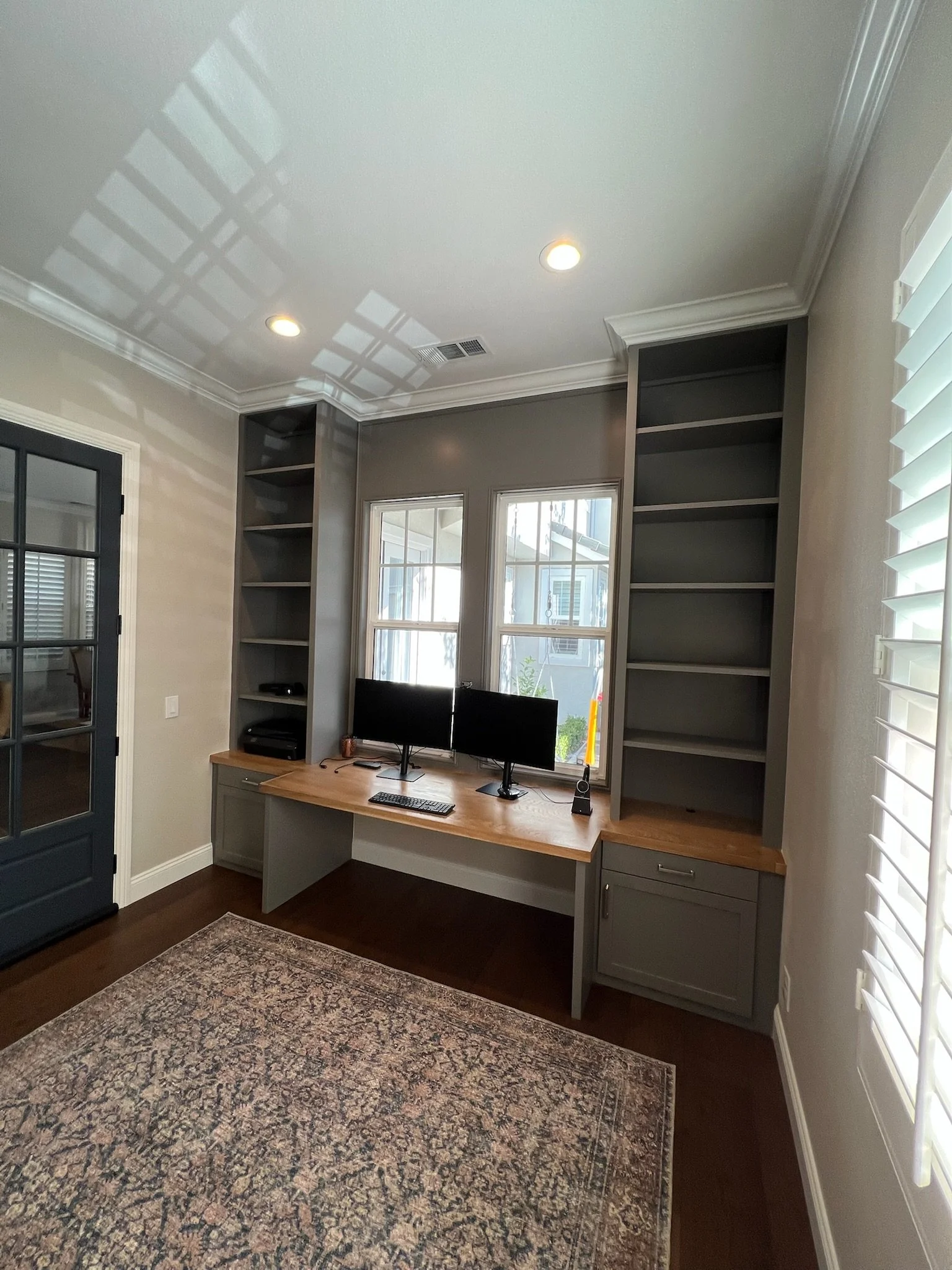 Empty built-in desk area with two computer monitors, situated in front of two windows with white blinds, flanked by tall gray bookshelves on either side, in a room with a patterned rug and a dark door.