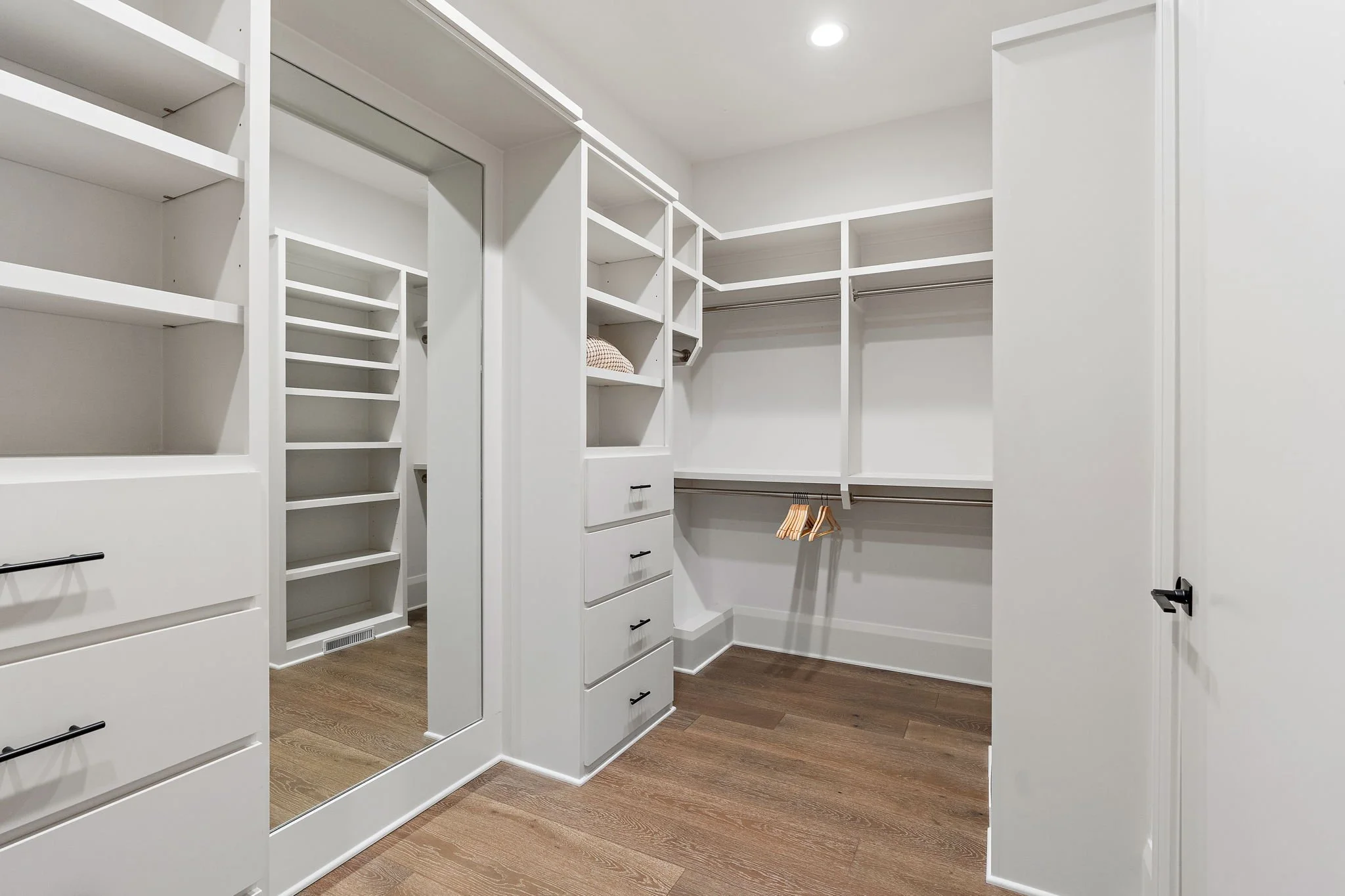 Empty walk-in closet with white shelves, drawers, and hanging rods, and hardwood flooring.