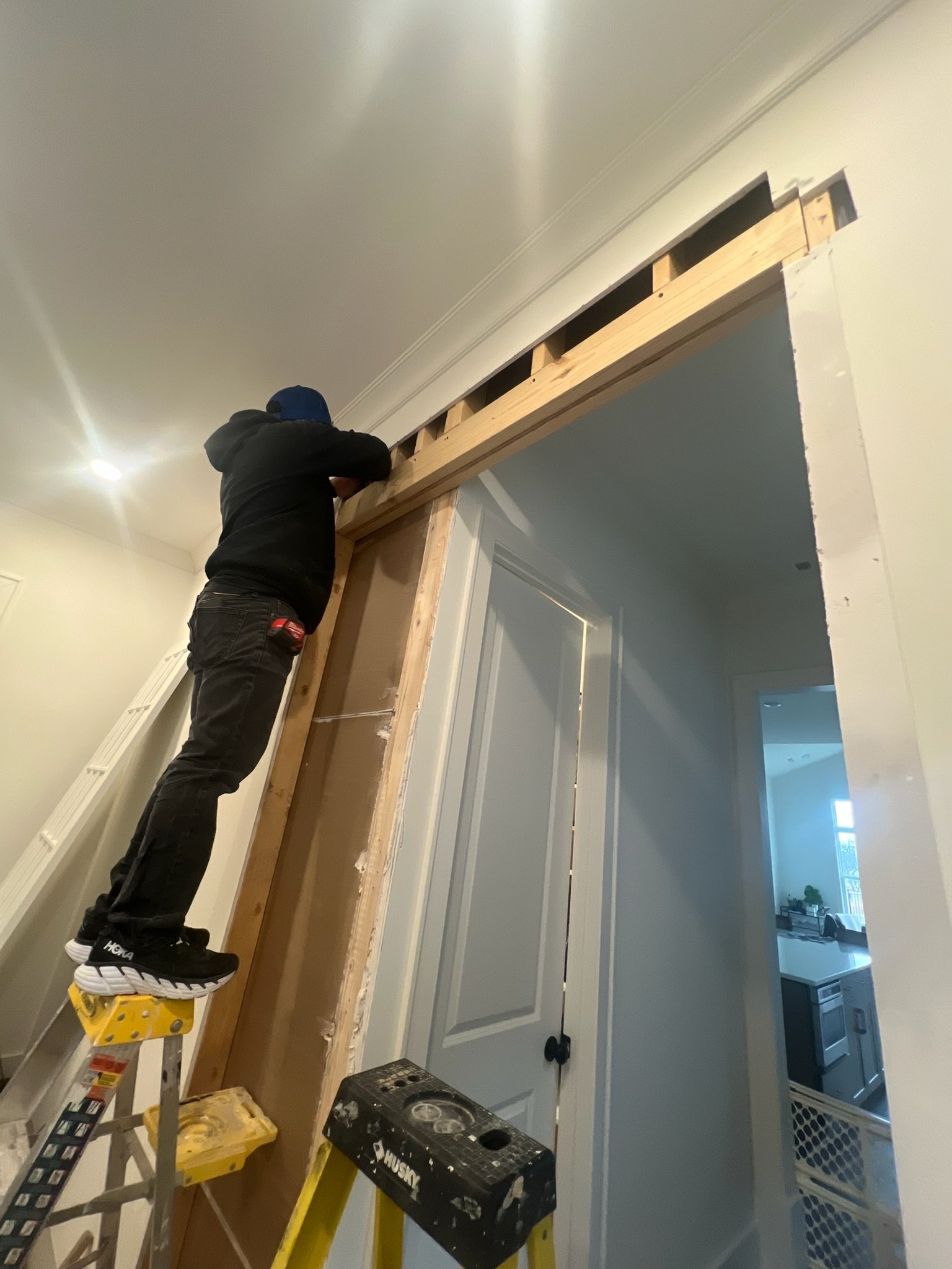 A person standing on a ladder working on a wooden door frame or curtain rod installation in an indoor room amidst a home renovation.