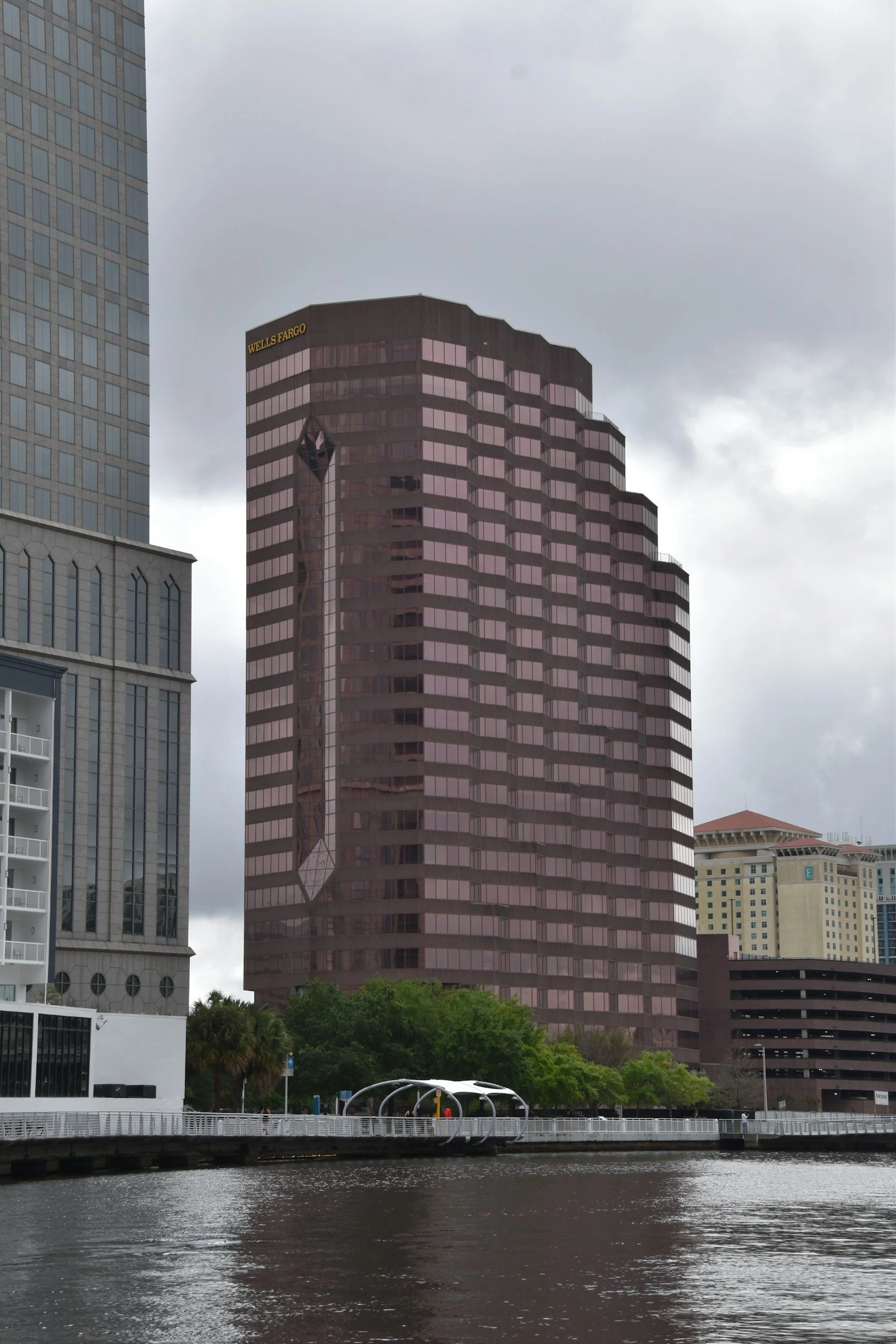 A cityscape featuring a pinkish-brown high-rise building with the Wells Fargo logo, adjacent modern buildings, a water body in the foreground, and a park with trees and a pathway along the water.