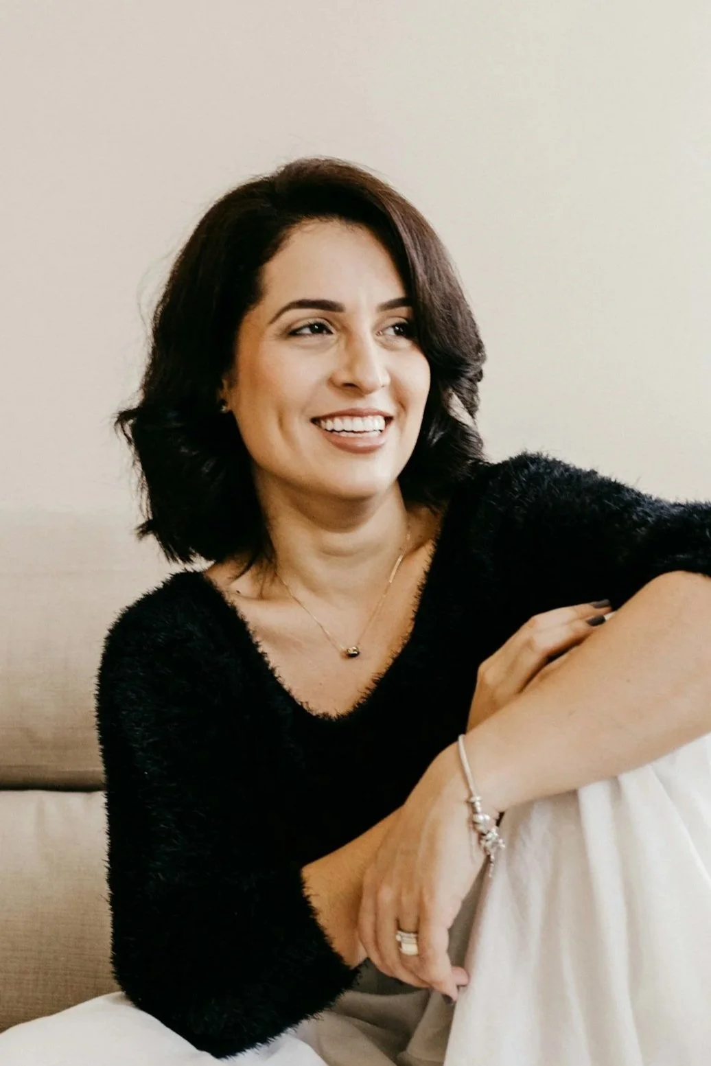 A woman with dark hair and a wide smile, wearing a black fuzzy sweater, sitting on a striped blanket on the floor with her legs crossed and holding her knees, in front of a beige sofa against a plain wall.
