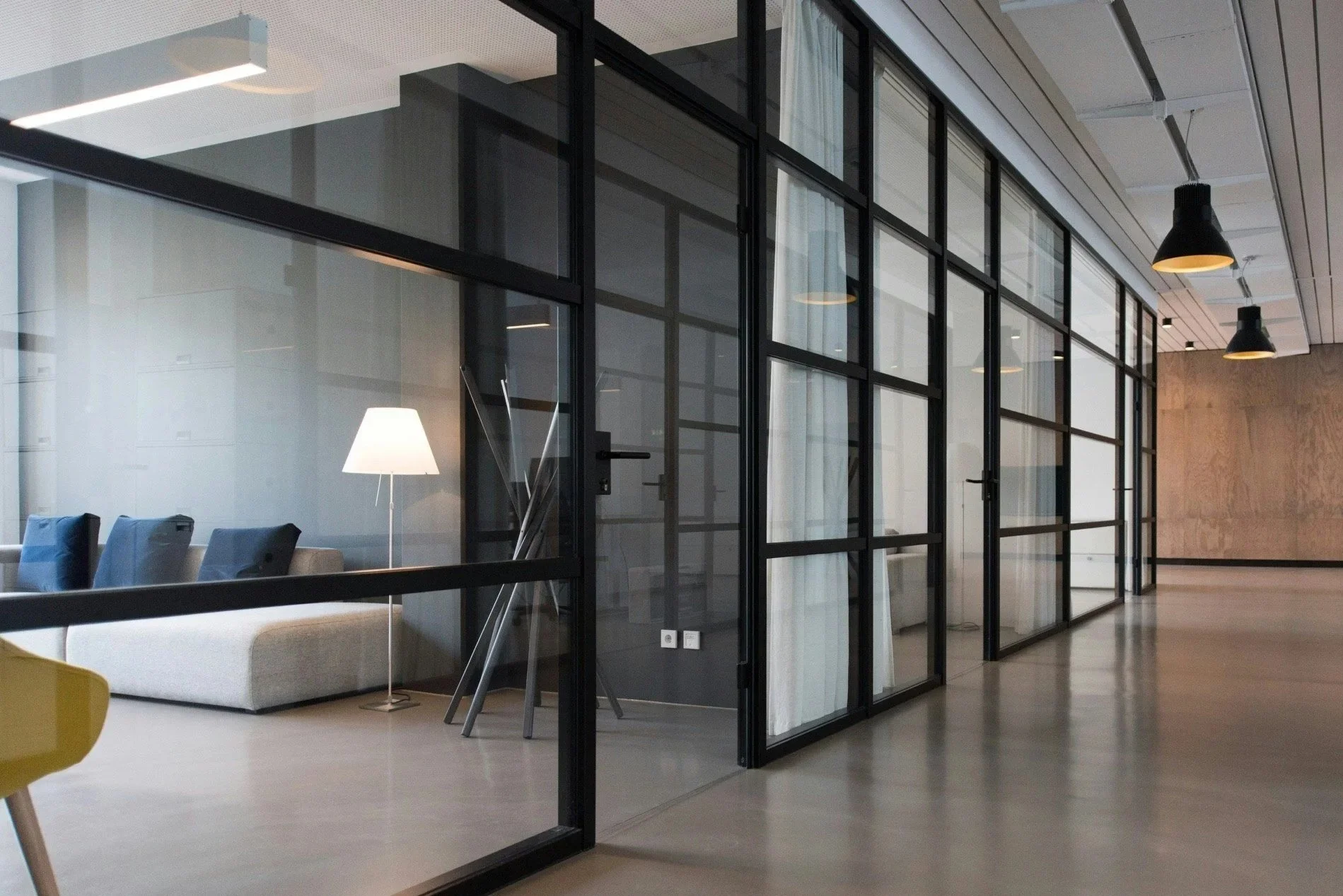 Modern office interior with glass partitions, a seating area with chairs and a floor lamp visible through the glass.