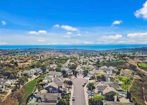 Aerial view of a suburban neighborhood near the coast with houses, streets, hills, and the ocean in the background under a blue sky with clouds.