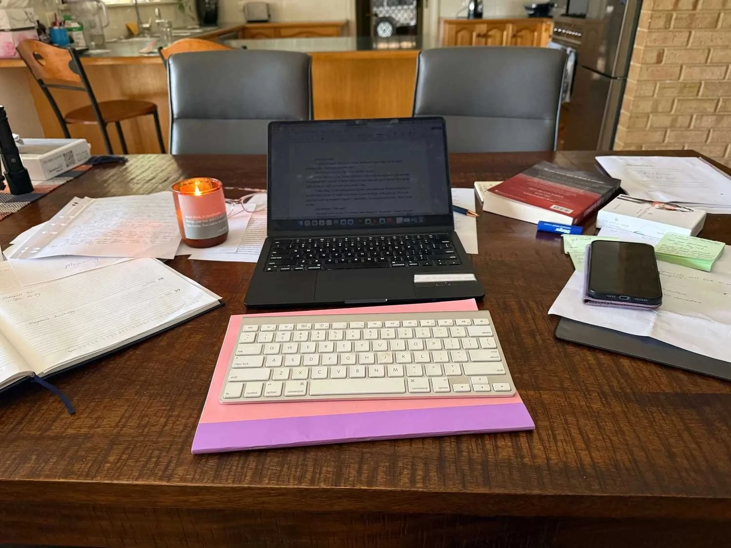 A laptop sits on a kitchen desk sournded by paper, a diary, a mobile phone, books, lists and a lit candle