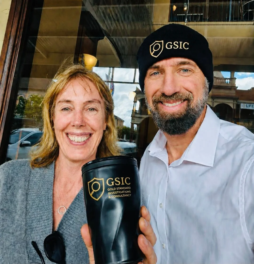 Fleur stands next to her friend, Andy, both are smiling. She is holding up a cup with his business logo on it, and he wears a hat with the same logo
