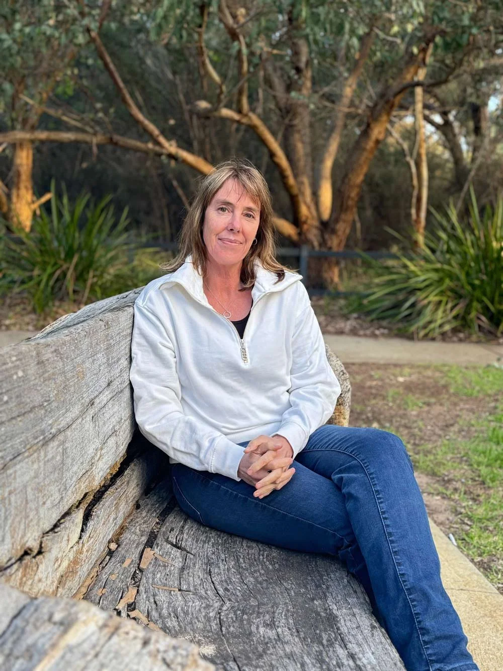 Fleur sits on a rustic wooden bench turning towards the camera, her legs are crossed with her hands clasped on her lap. Behind her is beautiful bush.