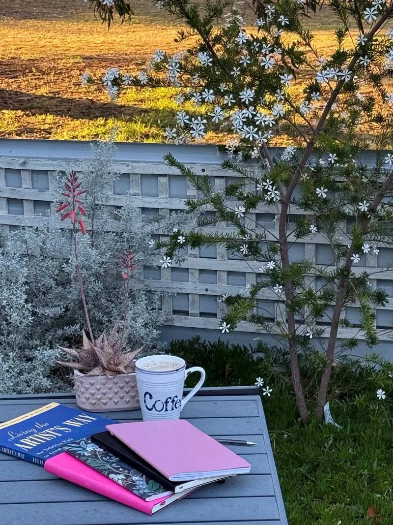 Four colourful diaries sit on a wooden table next to a coffee in a serene backyard setting