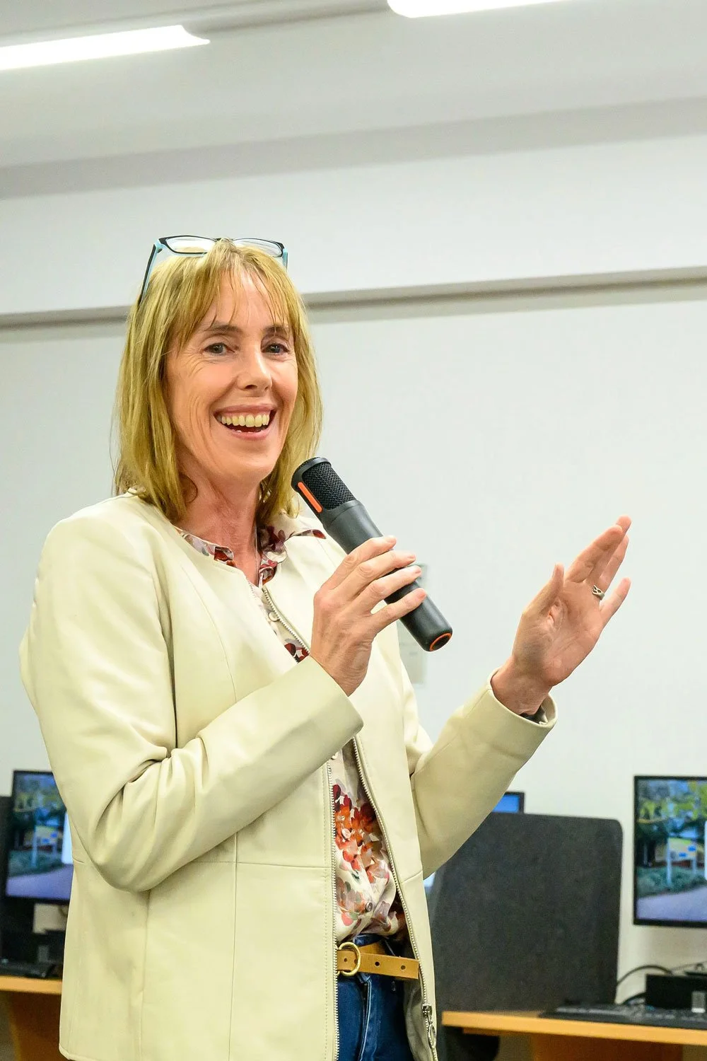 Fleur stands smiling and looking at the camera, she has a microphone in hand in the middle of telling a story to a crowd