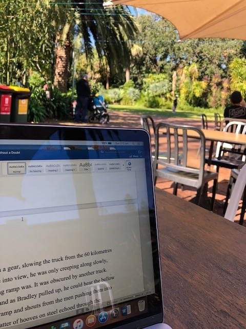 The top of a laptop on a wooden table under a shade. Lots of greenery in the background.