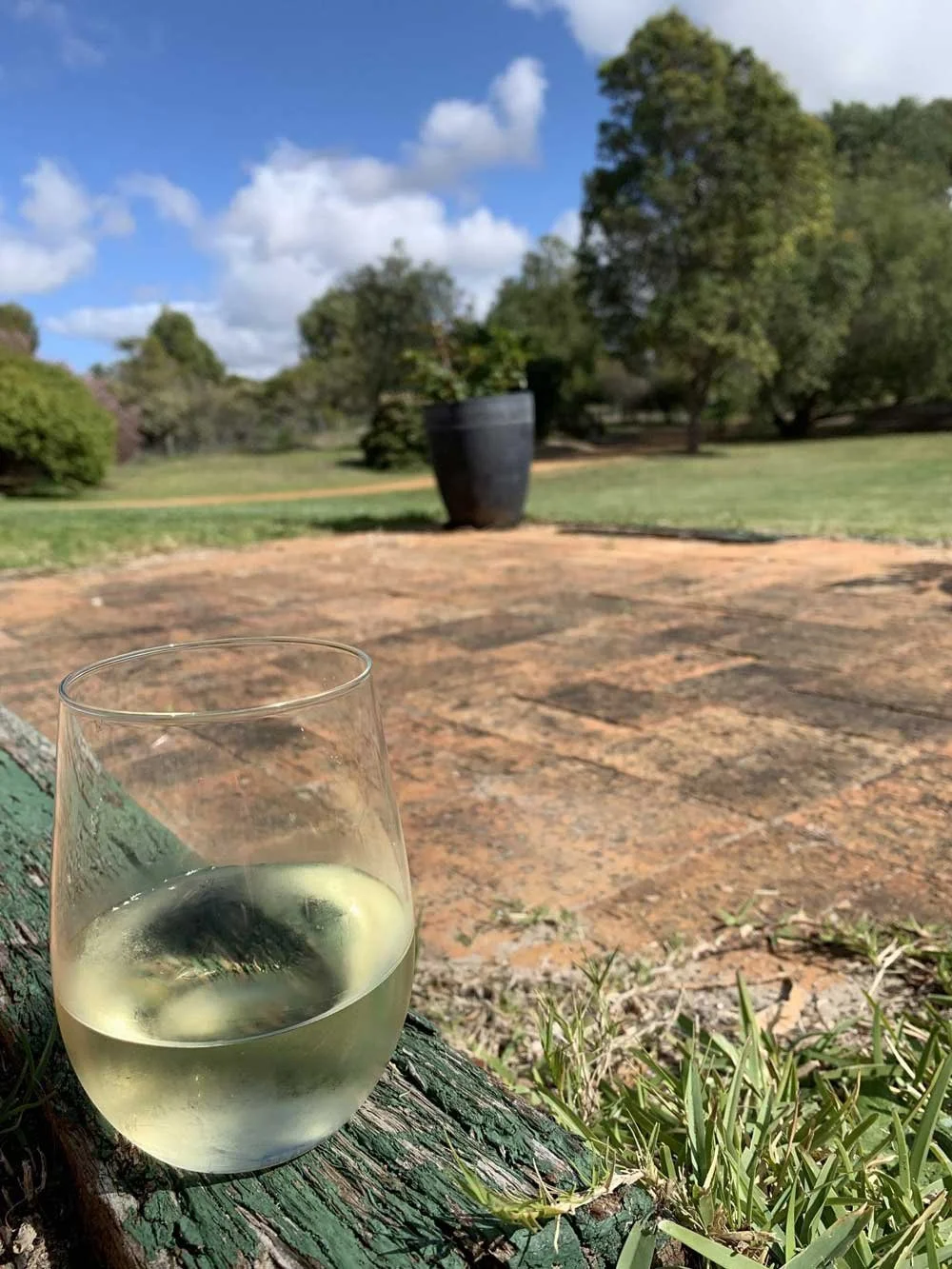 A stemless glass of white wine, sits on a wooden bench looking over a beautiful green yard