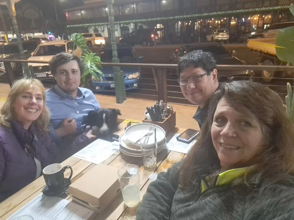 At a resturant table outside sit four people smiling at the camera