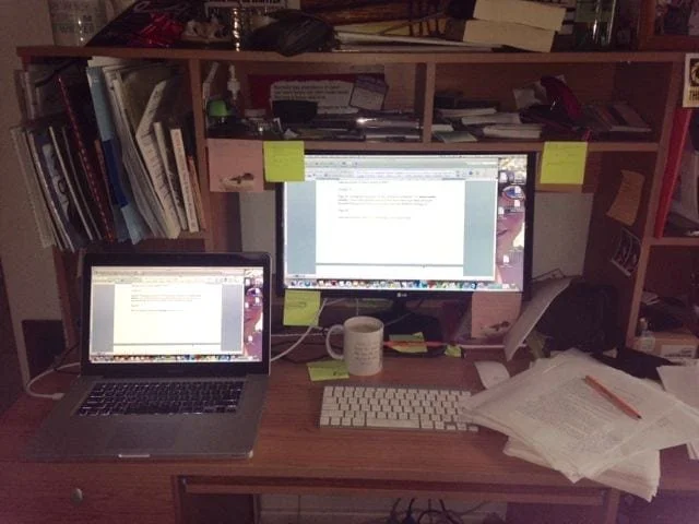 A busy desk. A desktop computer, a laptop and spread out pages of a manuscript with a pencil on top