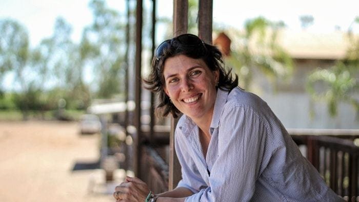 A woman stands smiling leaning over the balcony of a verandah. She has short brown hair and her sunglasses are on her head.