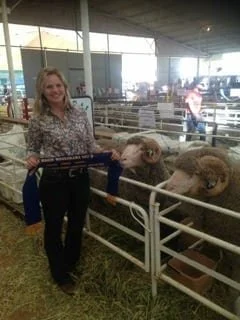A woman stands smiling holding a blue ribbon infront or beautiful rams in a shed