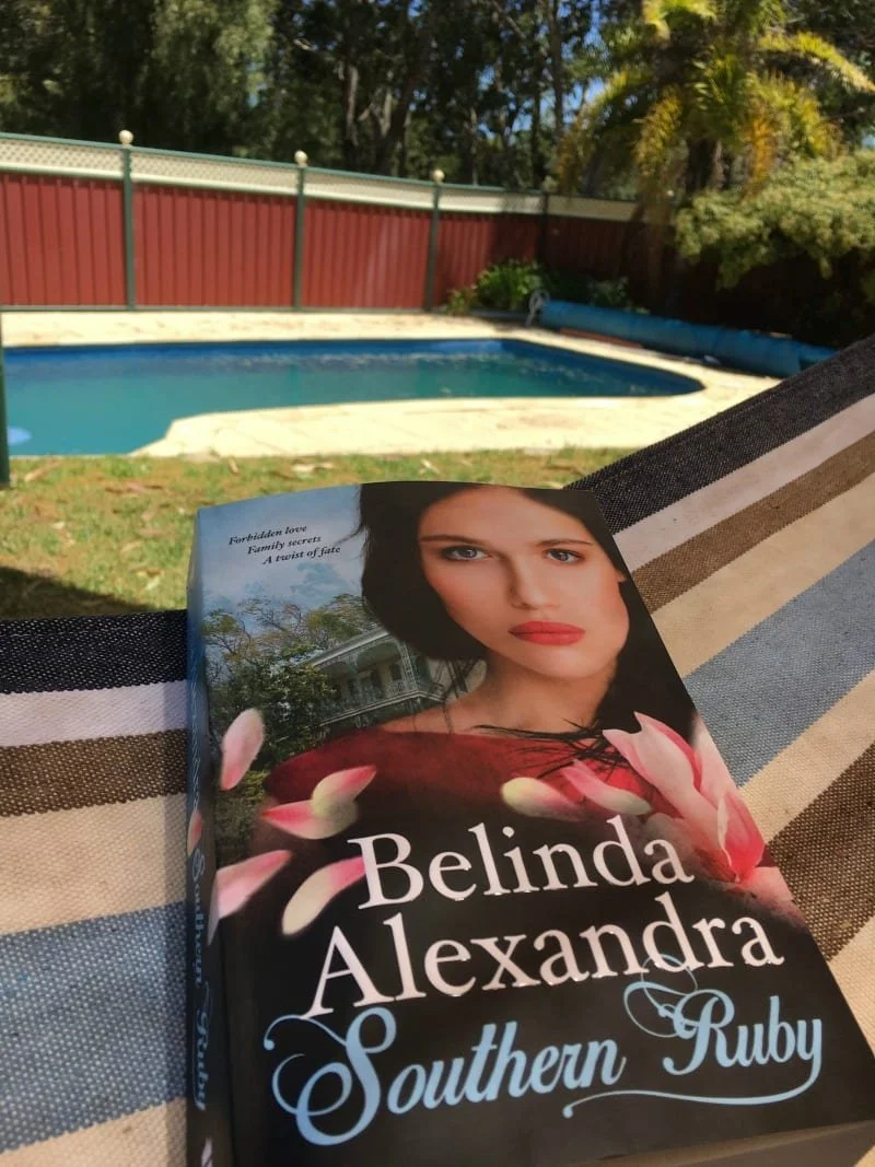 A book sits on a hammock. In the background, a pool. The book is Southern Ruby by Belinda Alexandra.