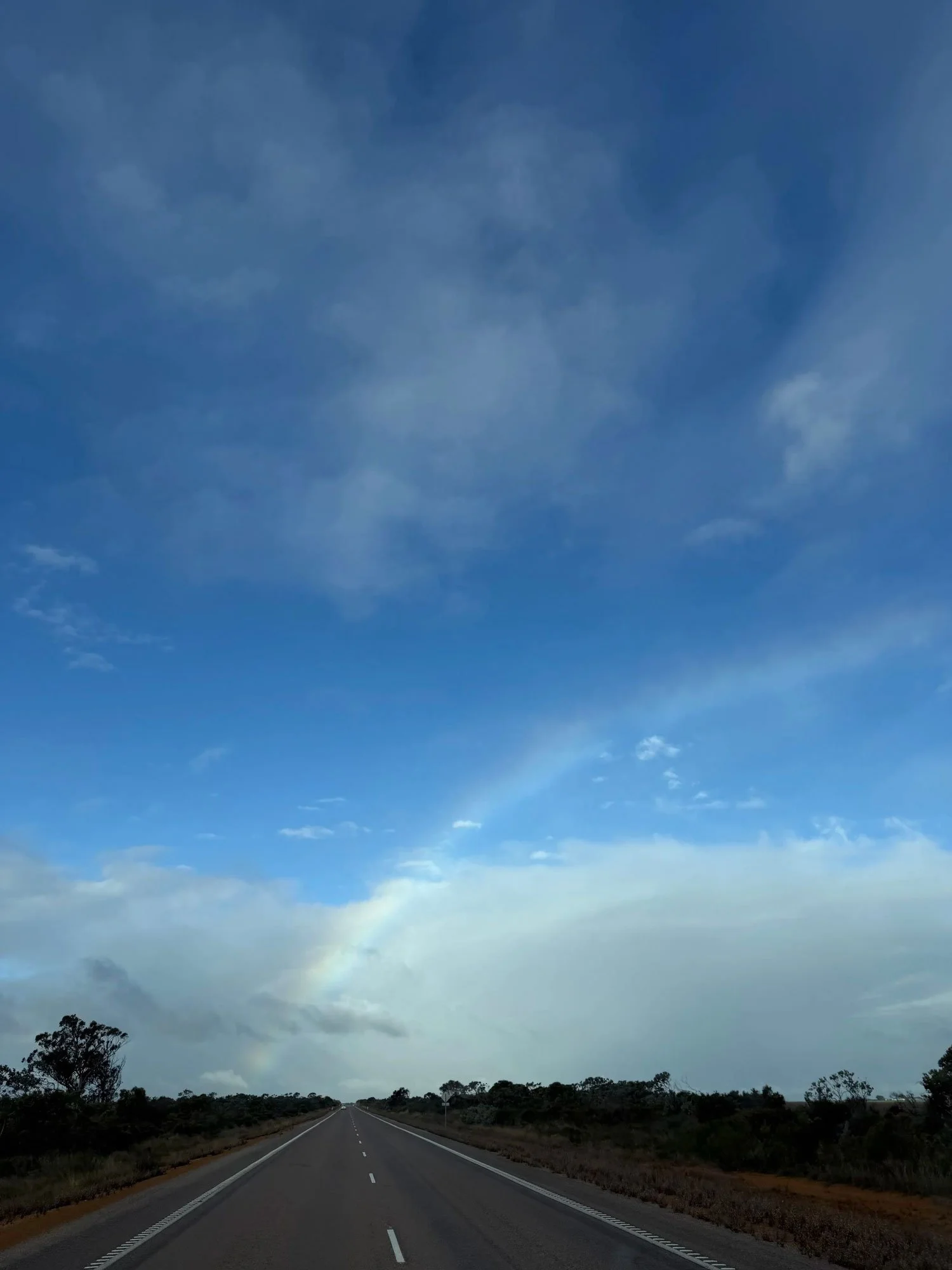 An open road with no cars, a blue sky and the HINT of a rainbow shining through clouds