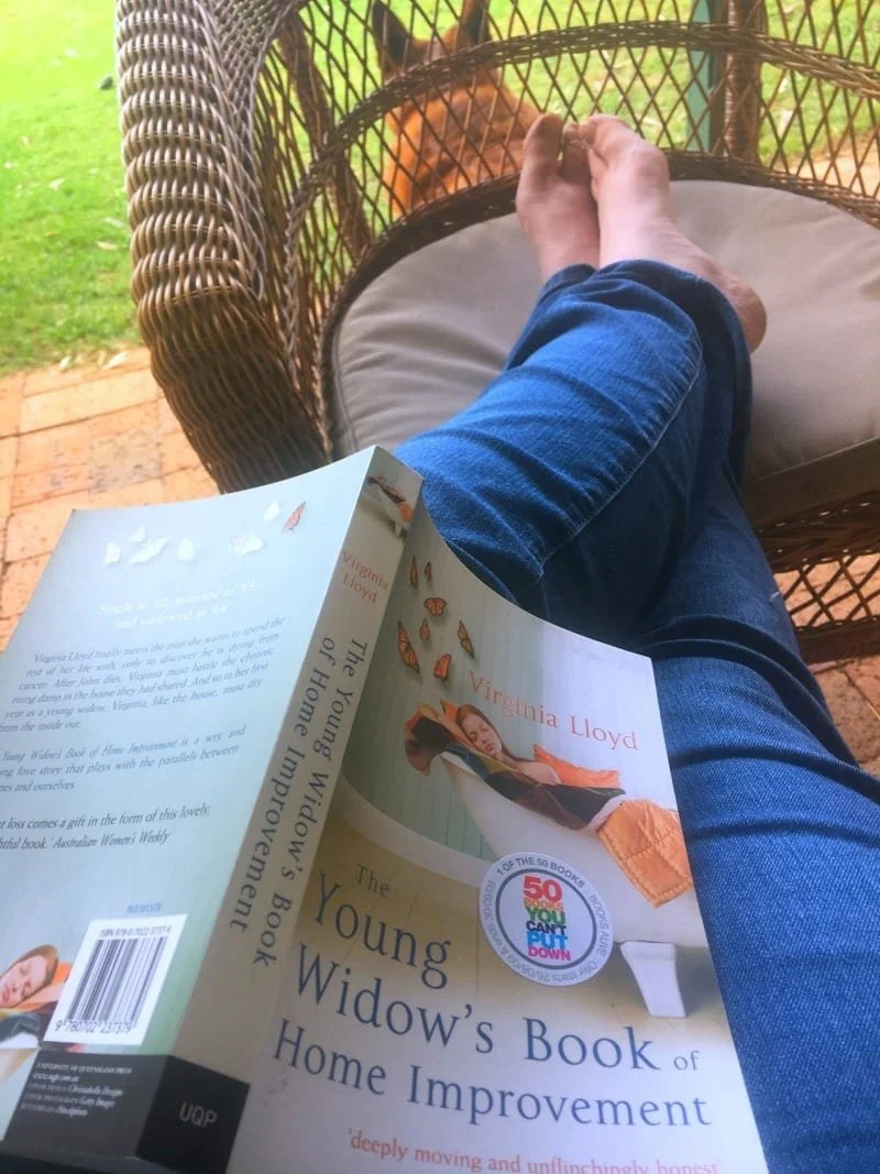 Feet up on a chair with a book over some legs. The book is The Young Widow's Book of Home Improvements.