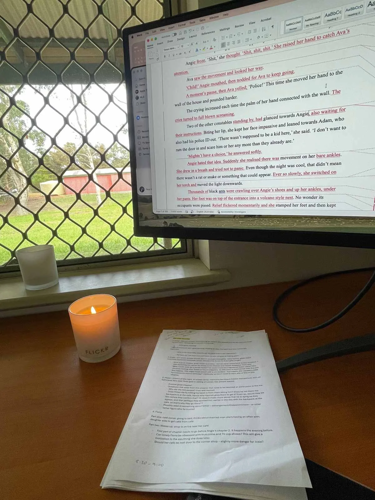 A computer screen shows a WIP of a book that is being edited. On the desk, a printed copy with lots of notes and highlighted sections. A lit candle sits on the desk too.