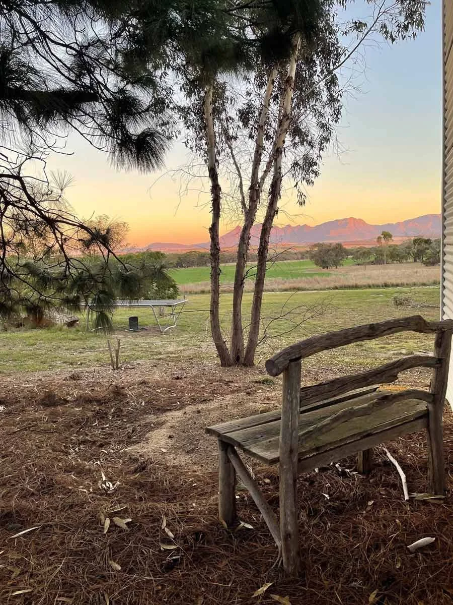A wooden bench looks over a yard with a trampoline. In the distance (not very far at all) you can see The Stirling Ranges