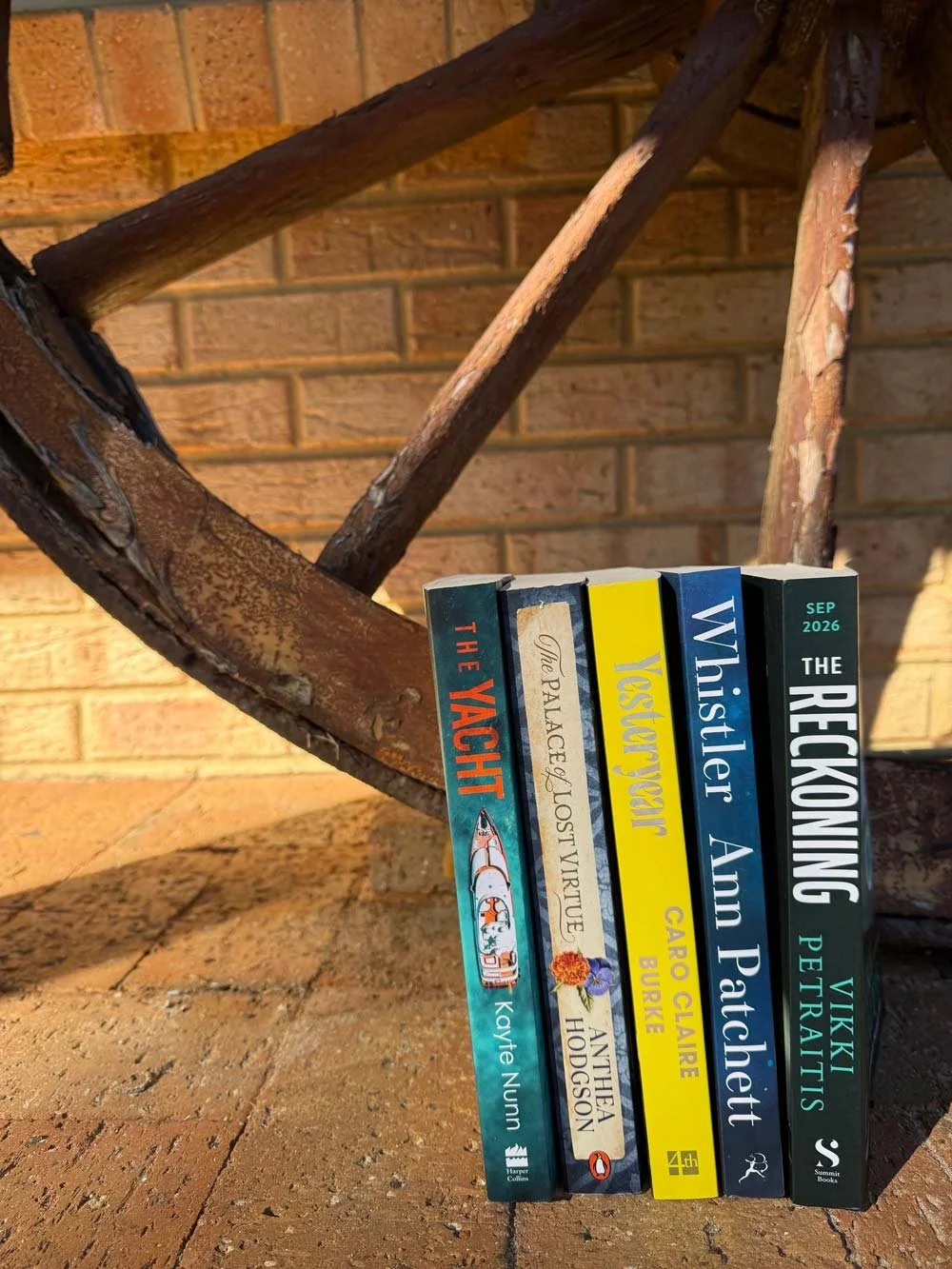 Four books together leaning against an old wagon wheel. The Yacht by Kayte Nunn, The Palace of Lost Virtue by Anthea Hodgson, Yesteryear by Caro Clair Burke, Whistler by Ann Patchett and The Reckoning by Vicki Petraitis.