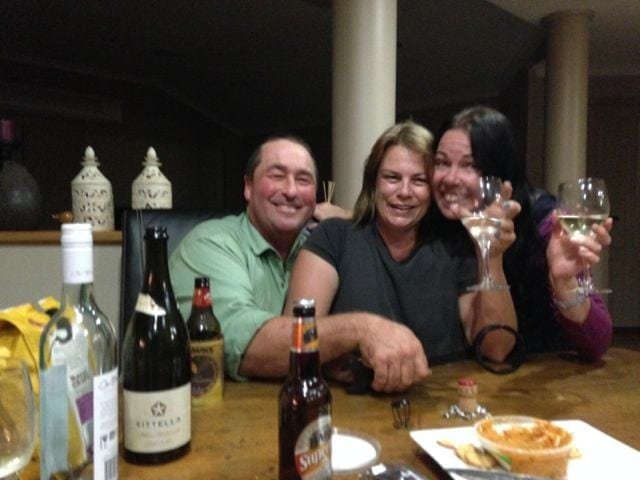 Two women and a man at table holding up glasses of wine smiling. The table has bottles and food on it