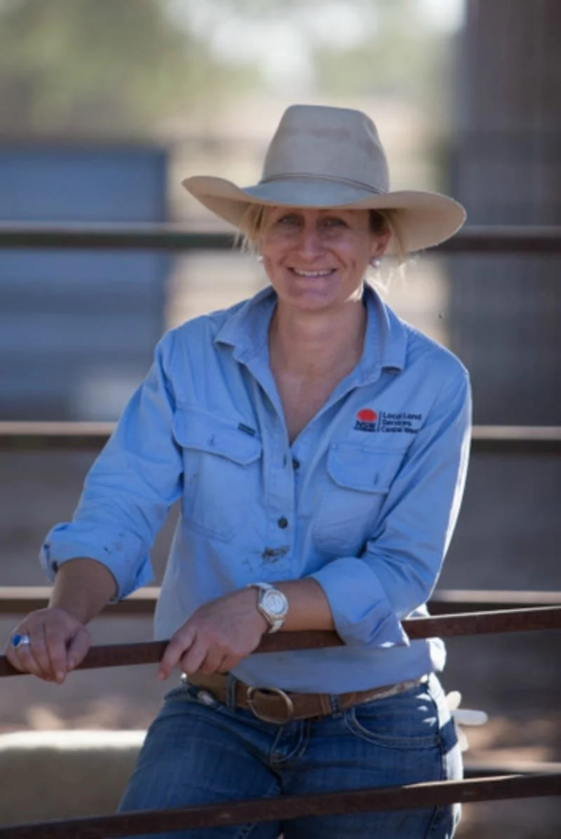 A woman smiles with beautitful dimples. She is in blue jeans, blue linen shirt and an akubra. She leans on a fence.