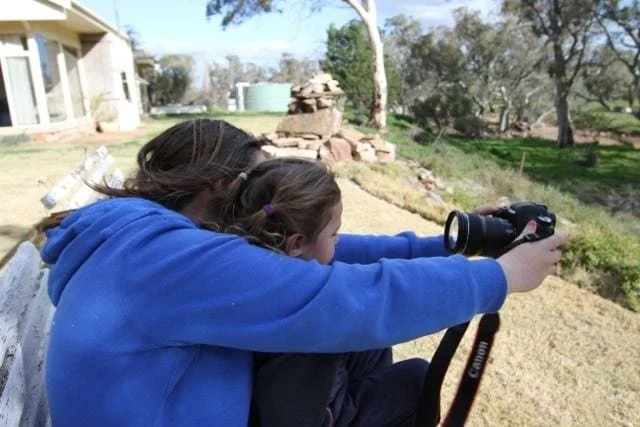 An older child envelops the younger one holding out a camera to take their picture
