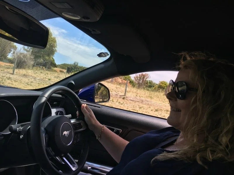 A woman smiling sitting in her car. The photo is taken from the passenger side of inside the car. The steering wheel has a horse in the middle ...