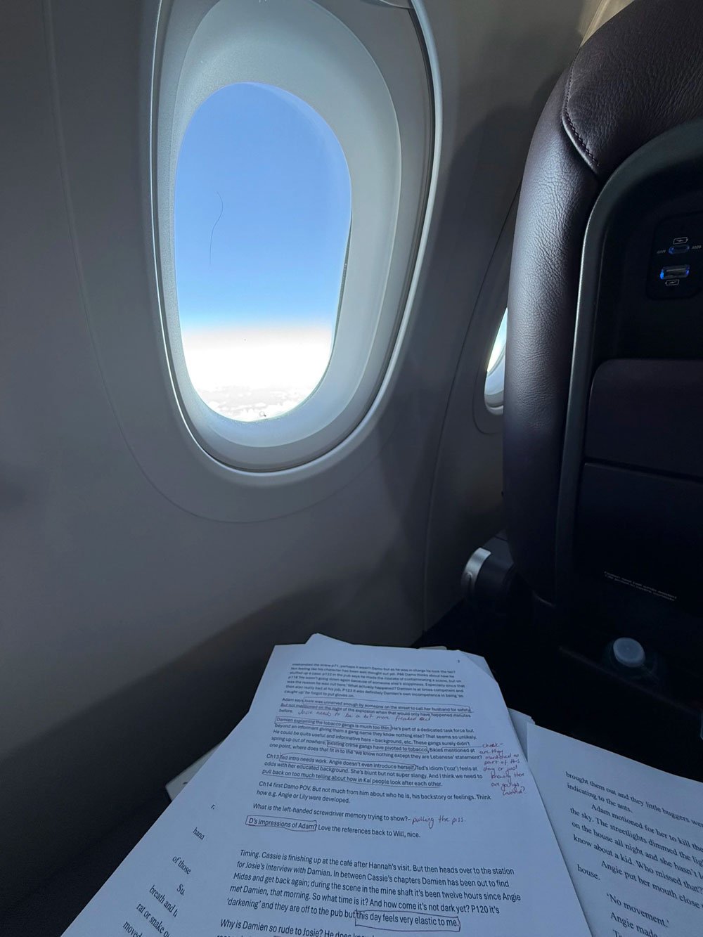 Looking out the window on an airplane. On the fold down desk are pages and pages of a printed out manuscript with mark up and editing notes on them in red.